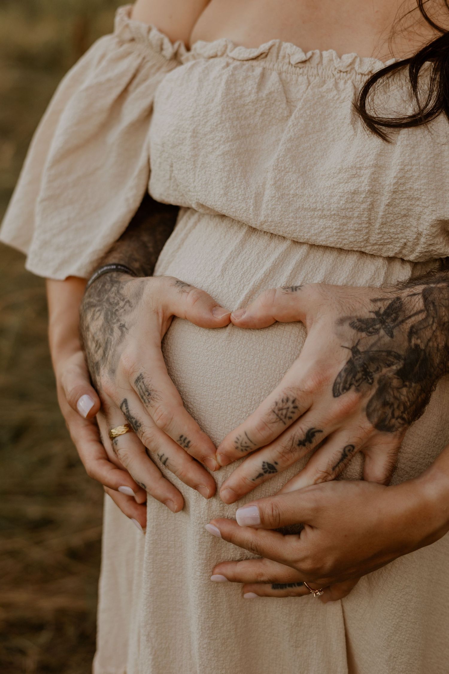 Pregnant woman in off-shoulder dress with tattooed hands forming heart shape over baby bump.