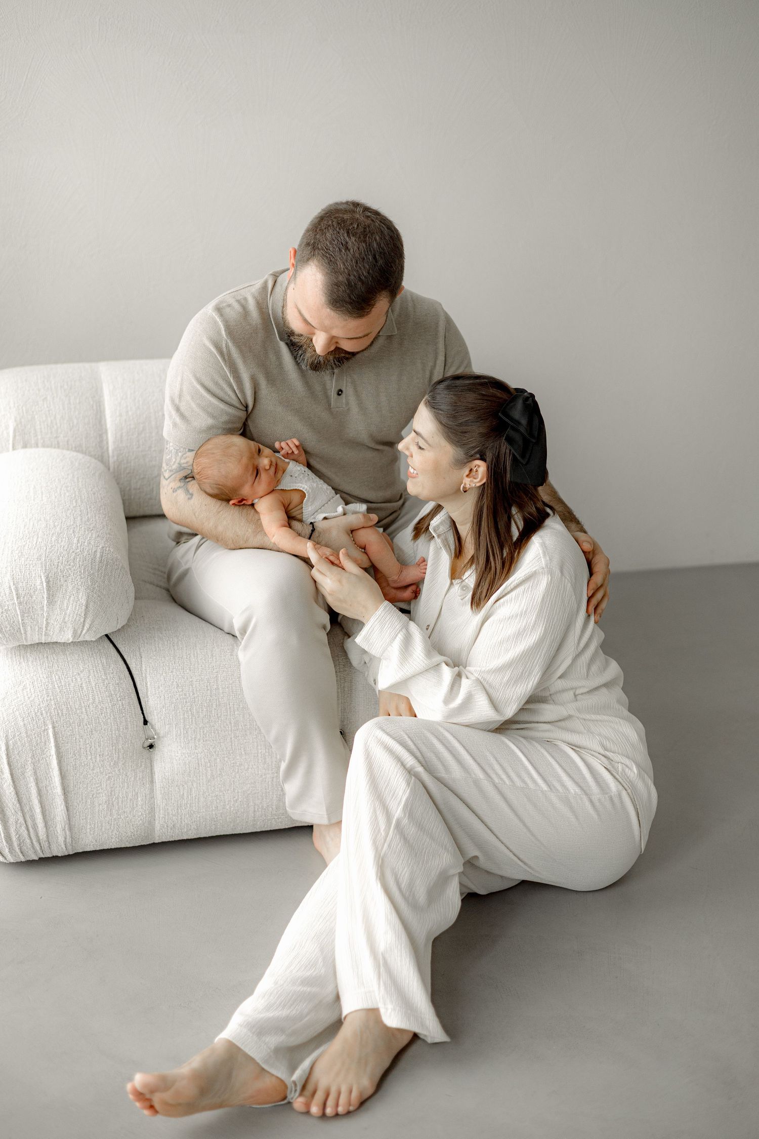 A couple sitting on a white sofa, lovingly looking at their newborn baby in a bright, minimalist room.