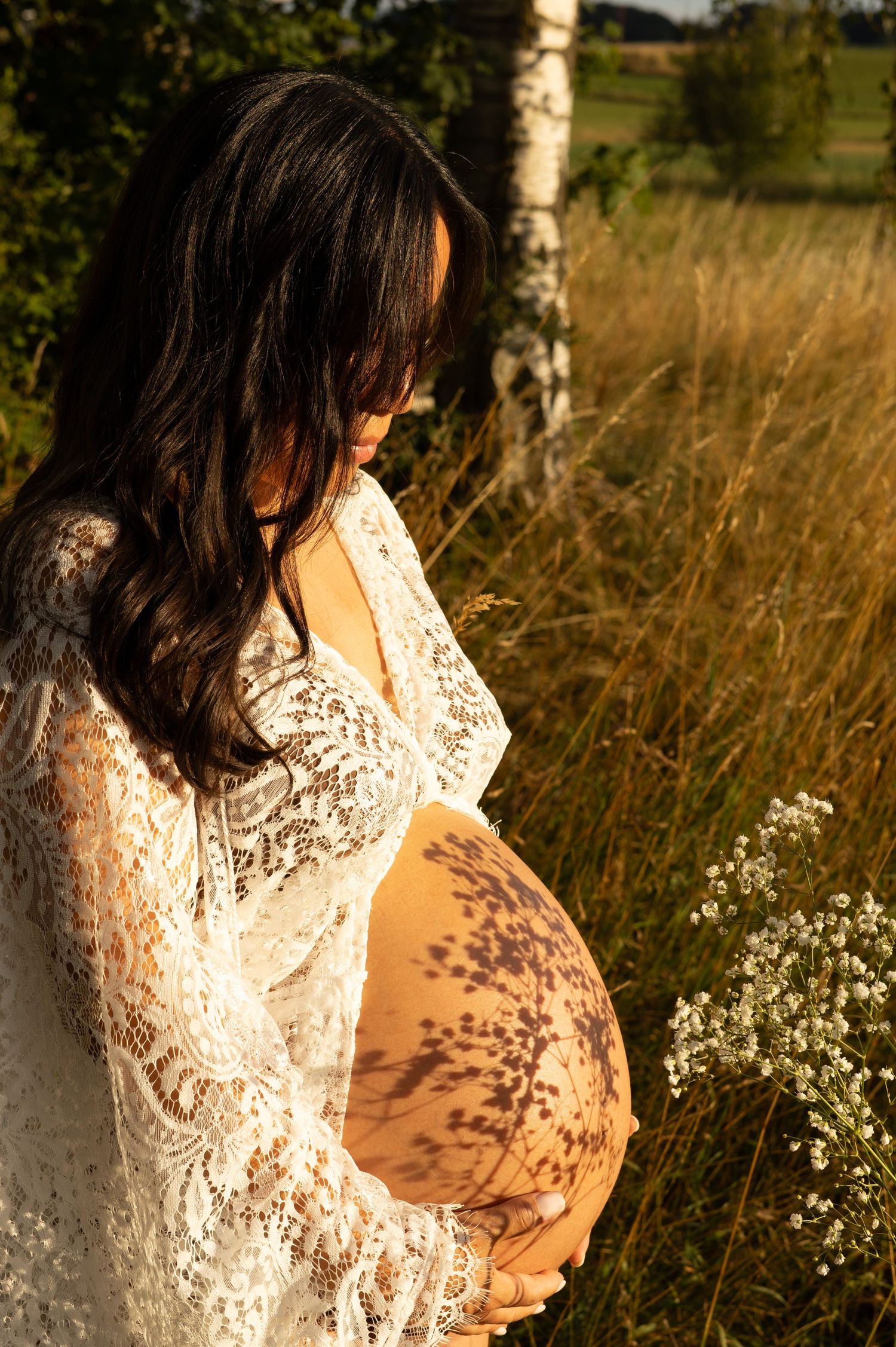 Pregnant woman in white lace robe cradling her baby bump outdoors in golden sunlight near birch trees and tall grass.