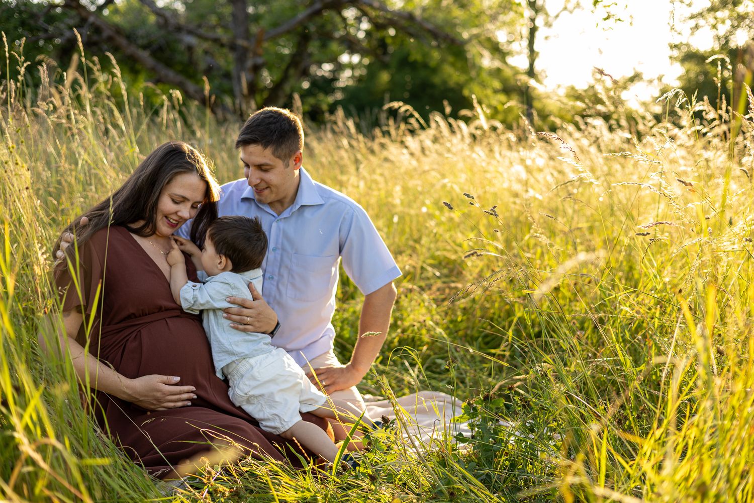 Pregnant woman and husband sit in sunny field with toddler, golden light highlighting their maternity family portrait.