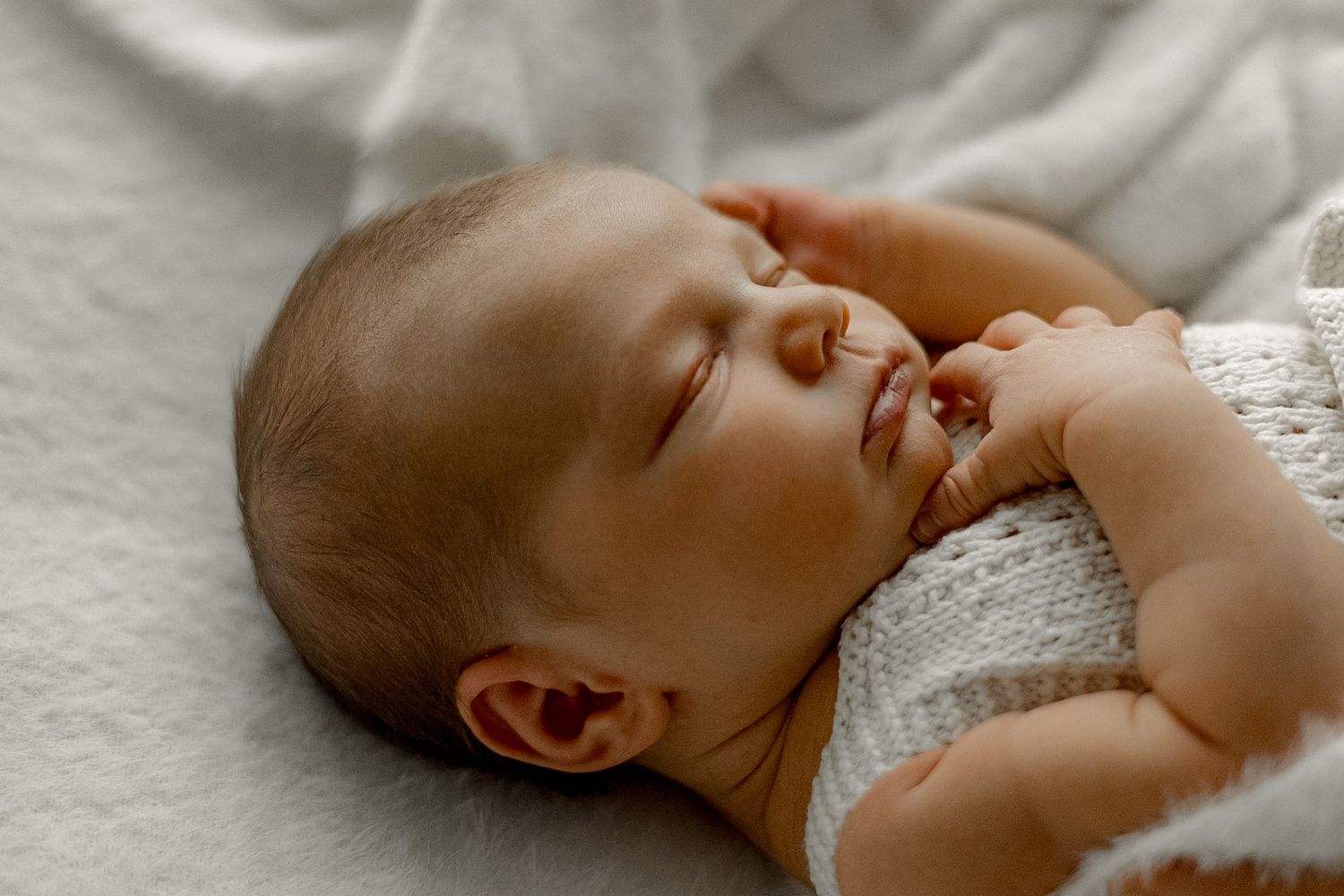 A peaceful newborn baby sleeping soundly, wearing a white knit outfit, with tiny hands near face on a soft blanket.