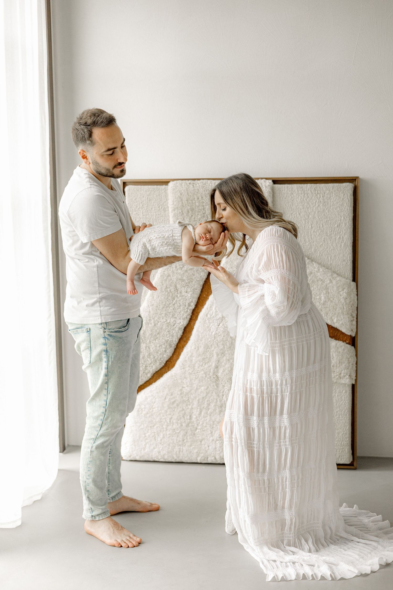 A couple in a bright studio admires their newborn baby, mother in white dress, father in casual attire.