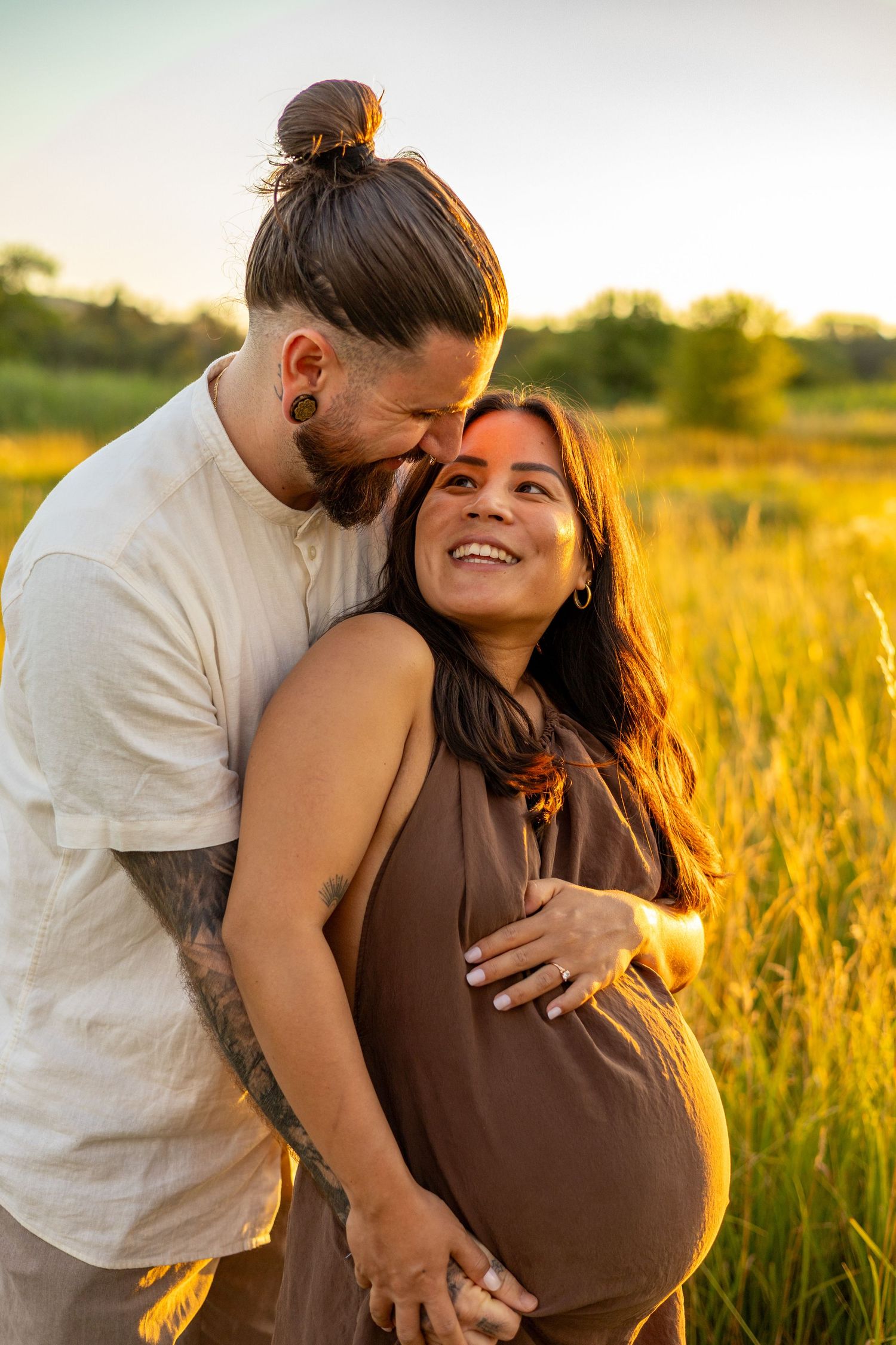 A happy pregnant couple embraces in a golden field at sunset, the man gently holding her baby bump.