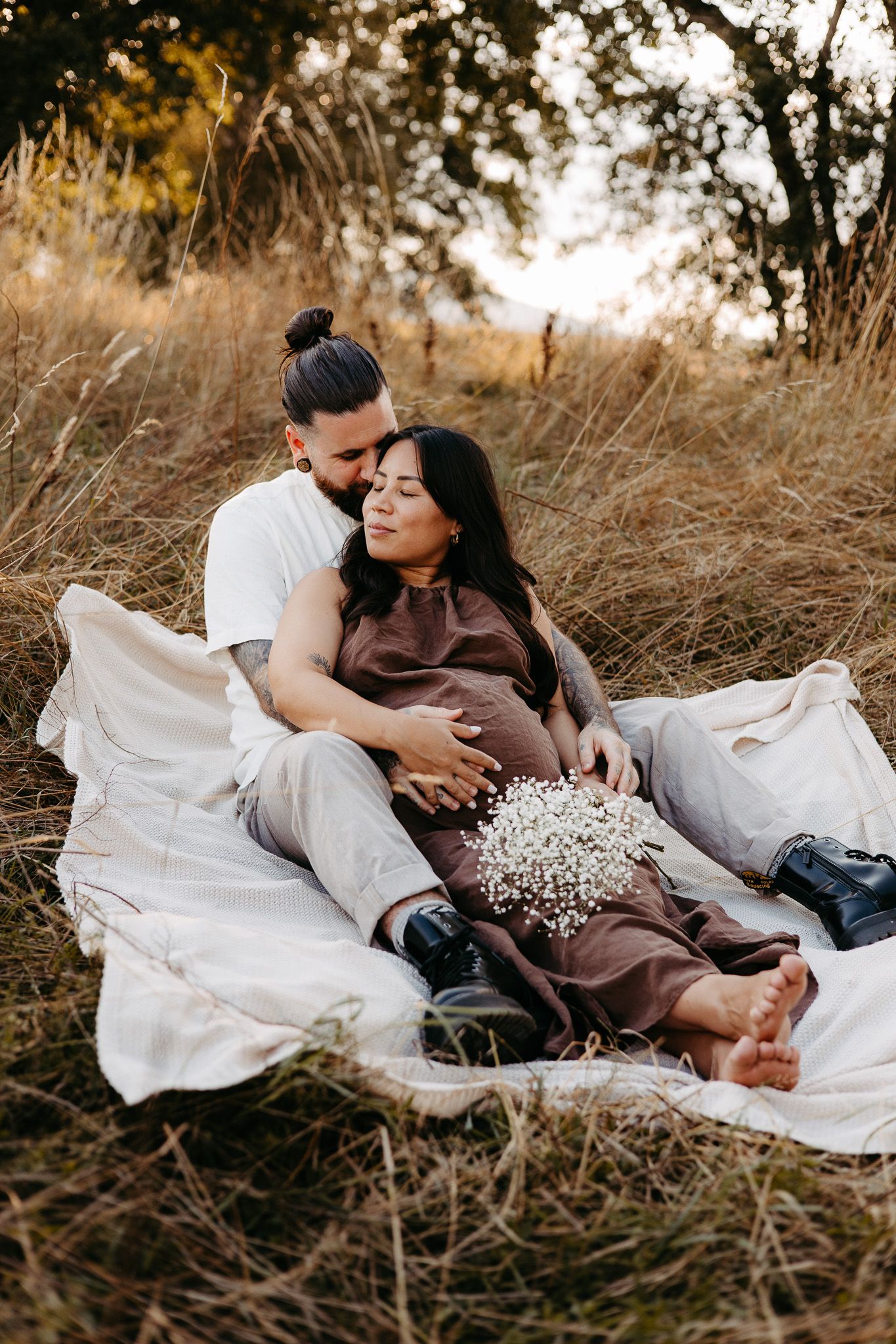 Couple sitting on white blanket in golden field, man embracing pregnant woman holding baby's breath flowers at sunset.