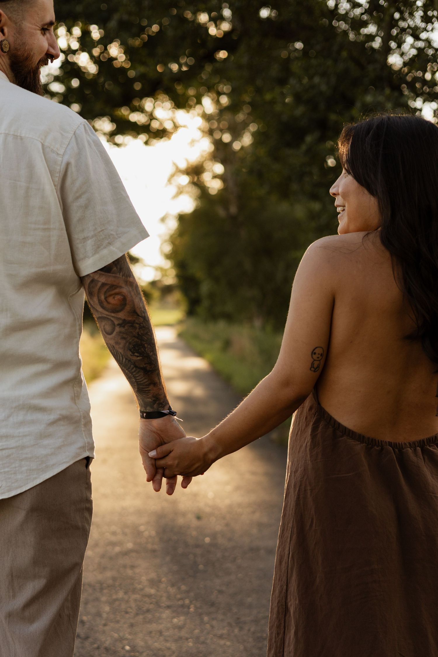 Tattooed couple holding hands on a sunlit path surrounded by trees at golden hour.