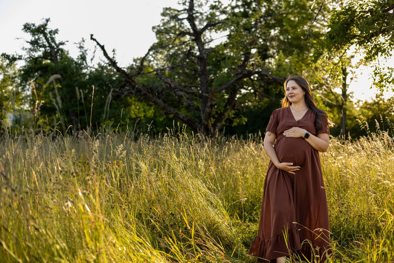 Pregnant woman in brown dress standing in tall grass field surrounded by lush green trees during golden hour.