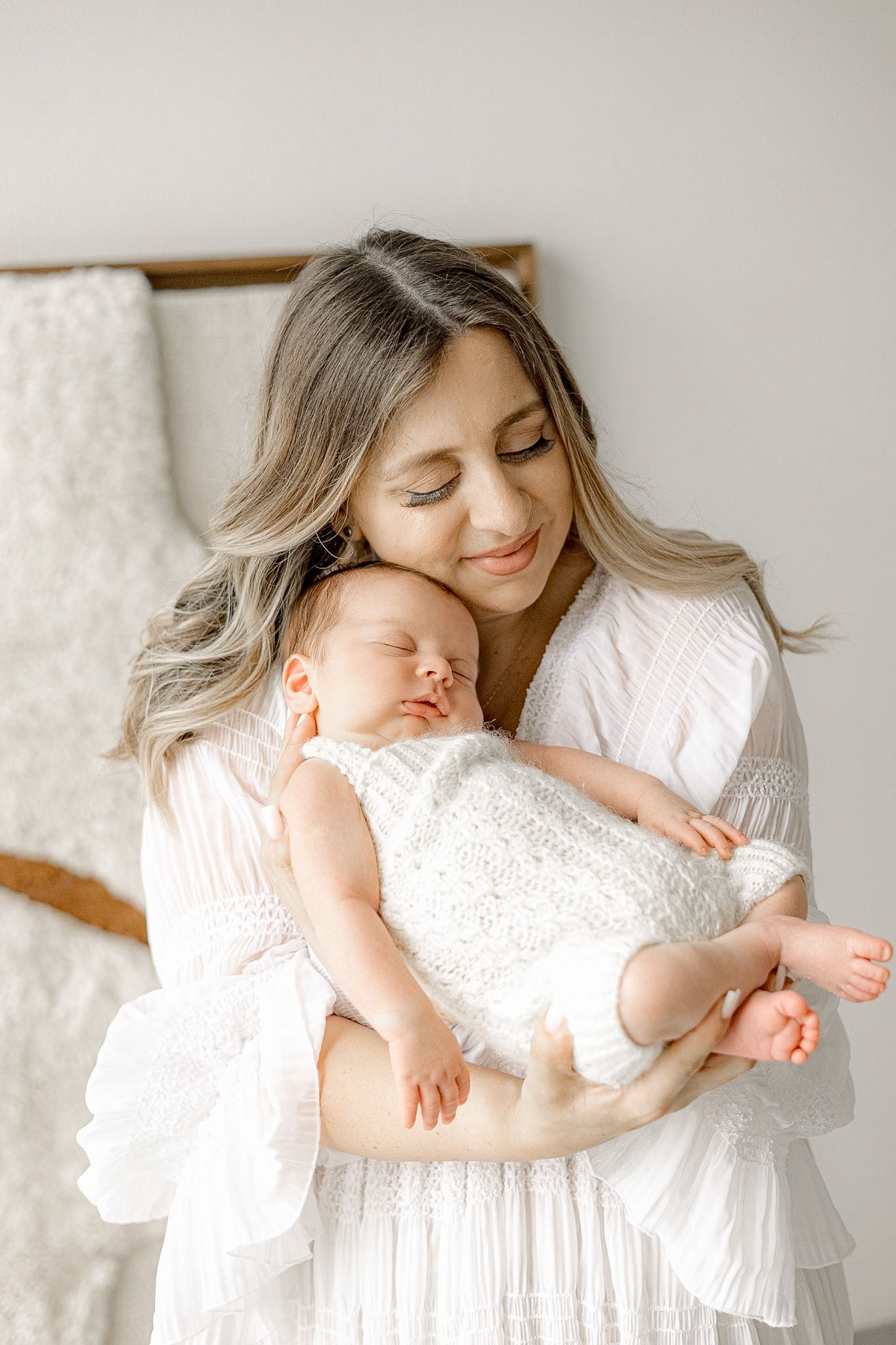 A mother in a white dress tenderly holds her sleeping newborn baby wrapped in a white lace outfit.