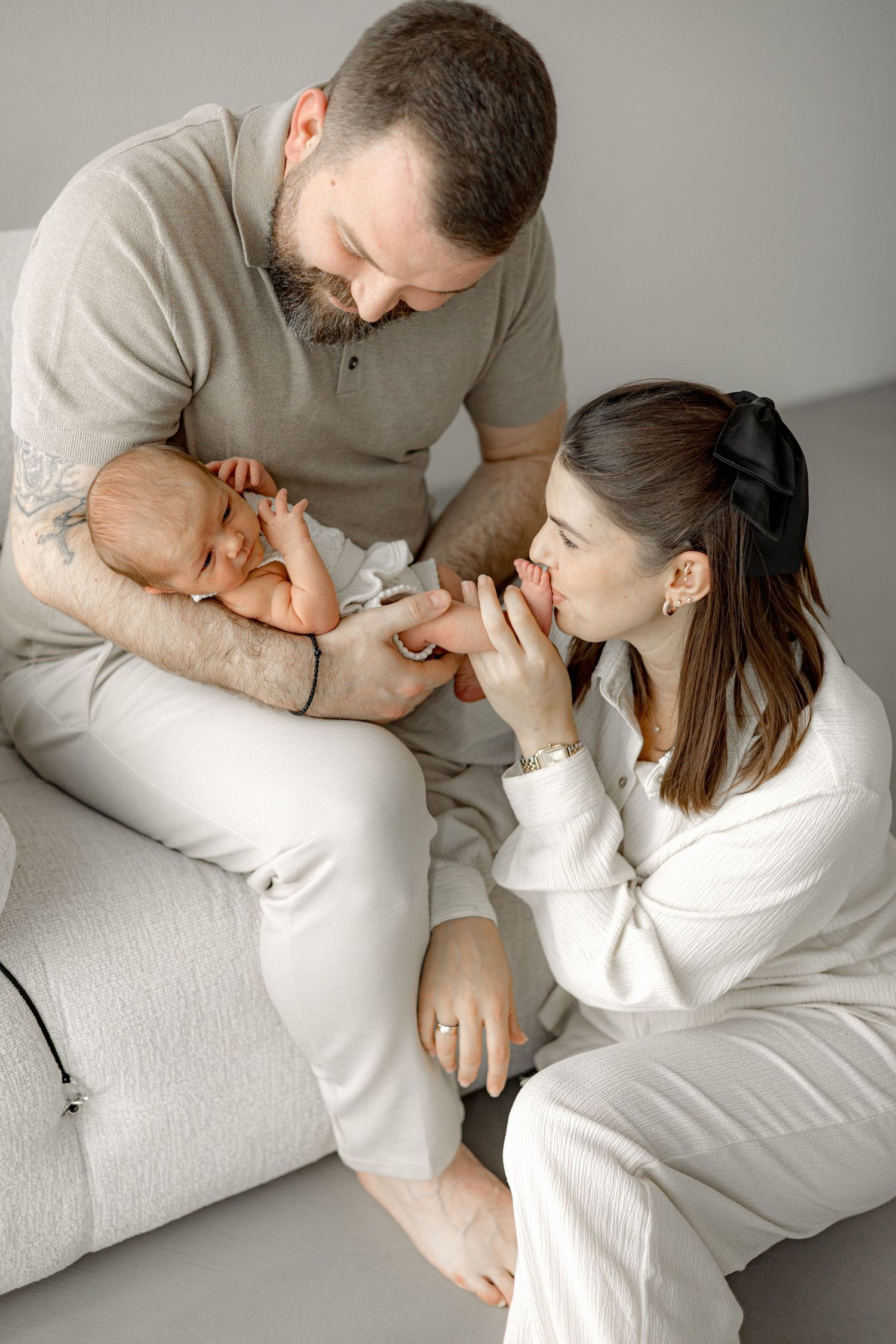 A bearded father holds a newborn baby while the mother kisses the infant's feet, sitting together on a light gray sofa.