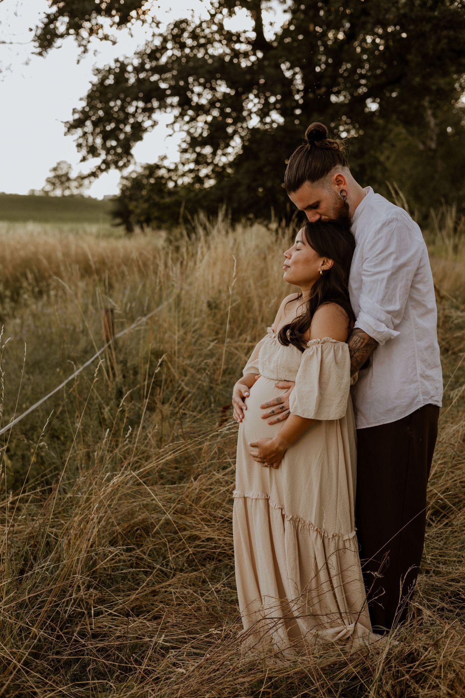 Couple sharing tender maternity moment in golden field, man embracing pregnant woman in beige dress at sunset.