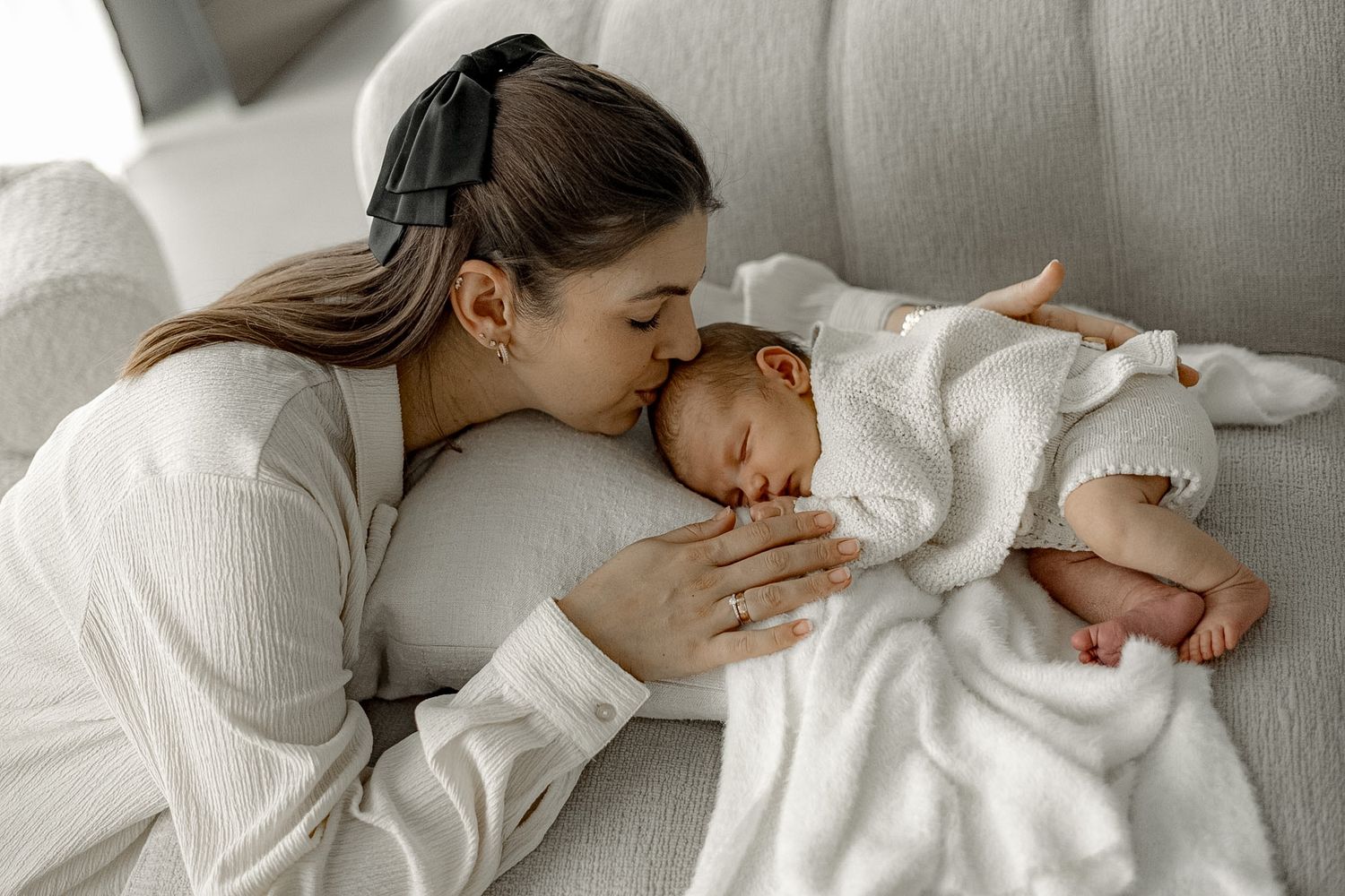 A young mother tenderly kisses her sleeping newborn baby lying on a white sofa covered in a soft blanket.