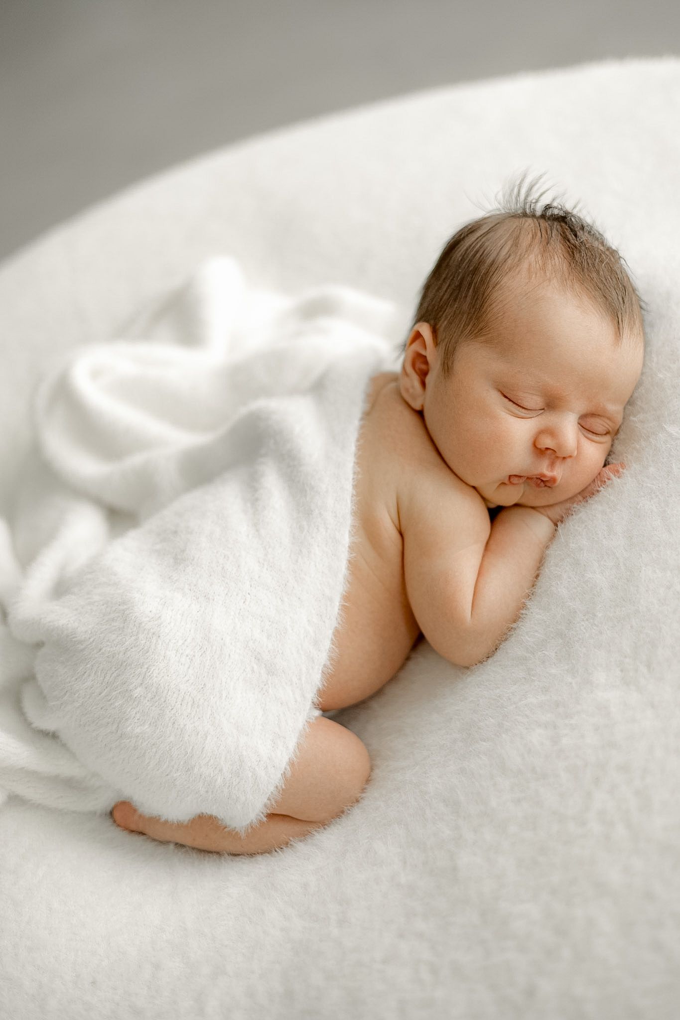 Peaceful newborn baby sleeping curled up on white fluffy blanket in soft natural light photography.