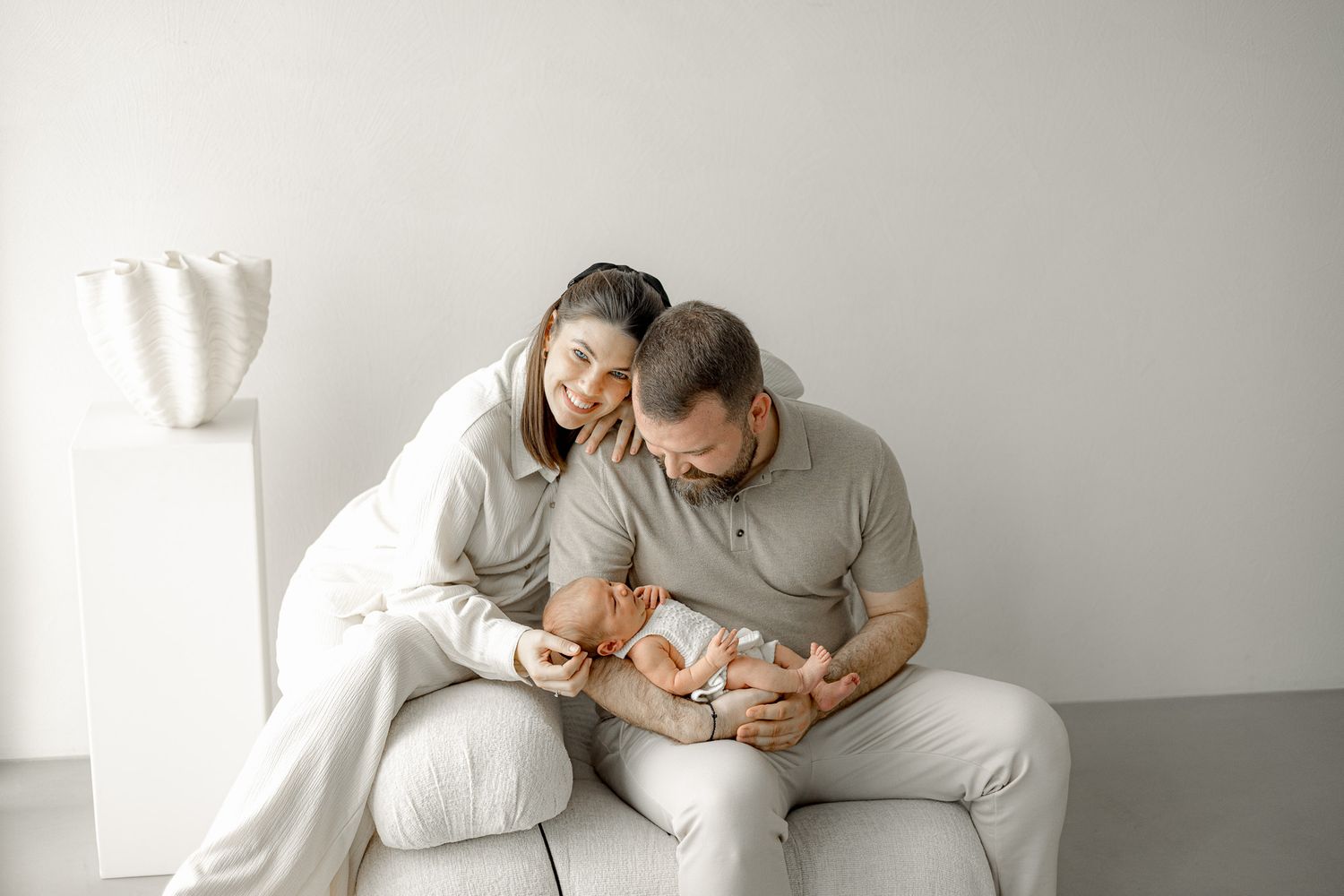 Happy couple sitting on white sofa admiring their newborn baby in a bright minimalist studio setting.