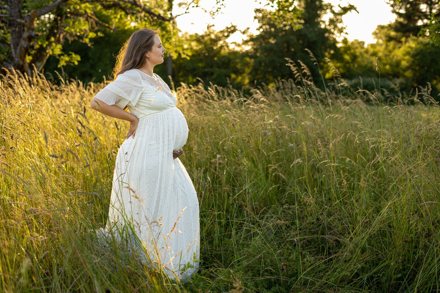 Pregnant woman in white dress standing in golden tall grass field at sunset, cradling baby bump.