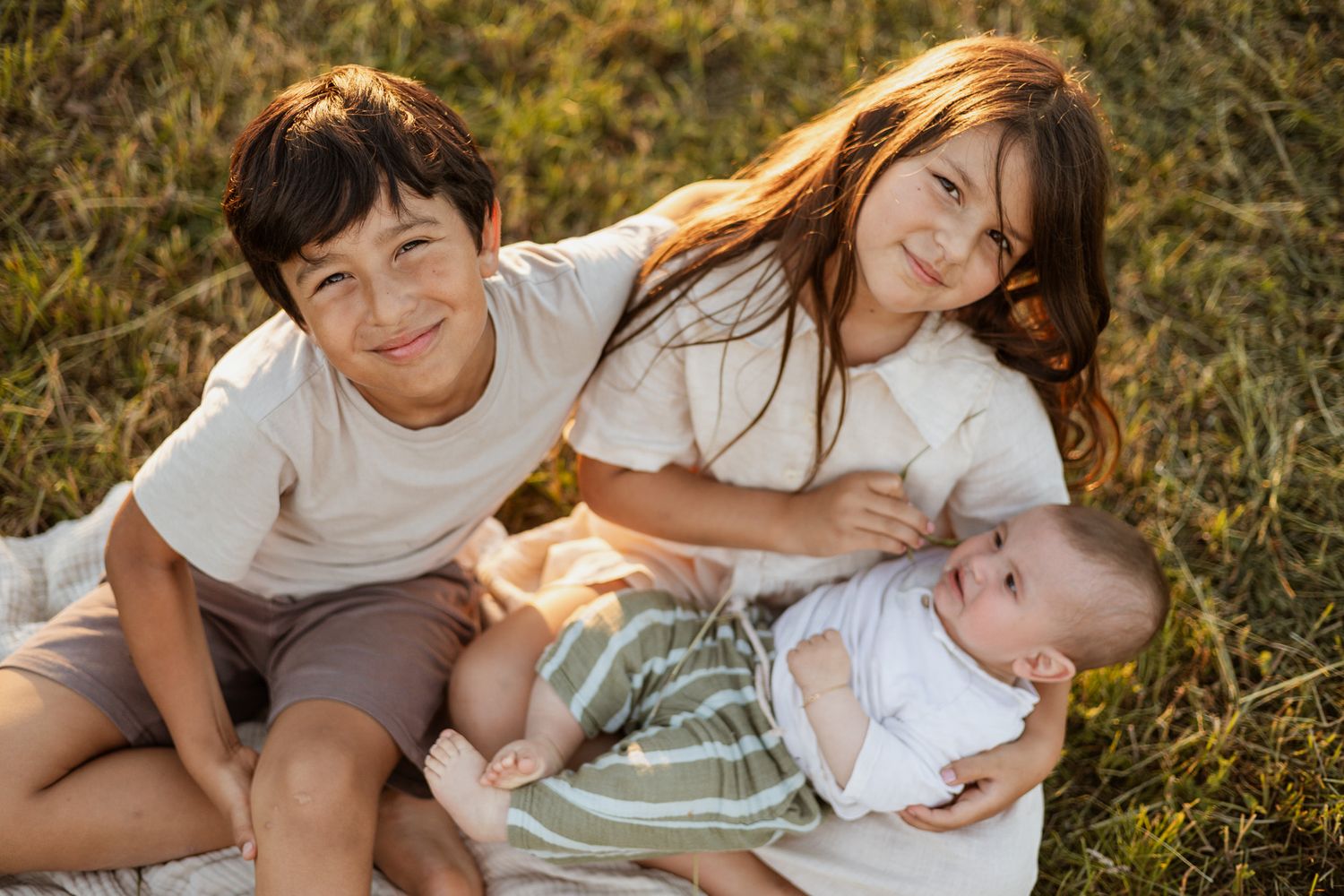 Two children and a baby sibling sit together on a blanket in a grassy field, smiling warmly in soft natural light.