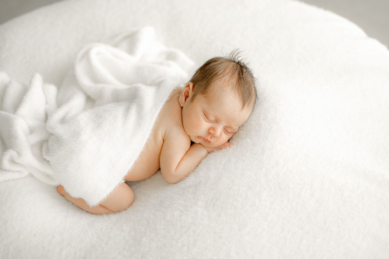 Peaceful newborn baby sleeping on a soft white blanket, curled up in a cozy position during a professional newborn photo session.