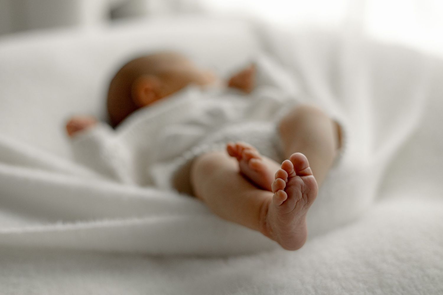 Newborn baby sleeping peacefully on white blanket with tiny feet in focus, soft natural light photography.