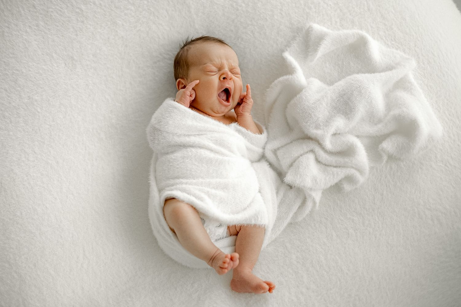 Newborn baby yawning while swaddled in a white blanket, lying on a soft white surface.