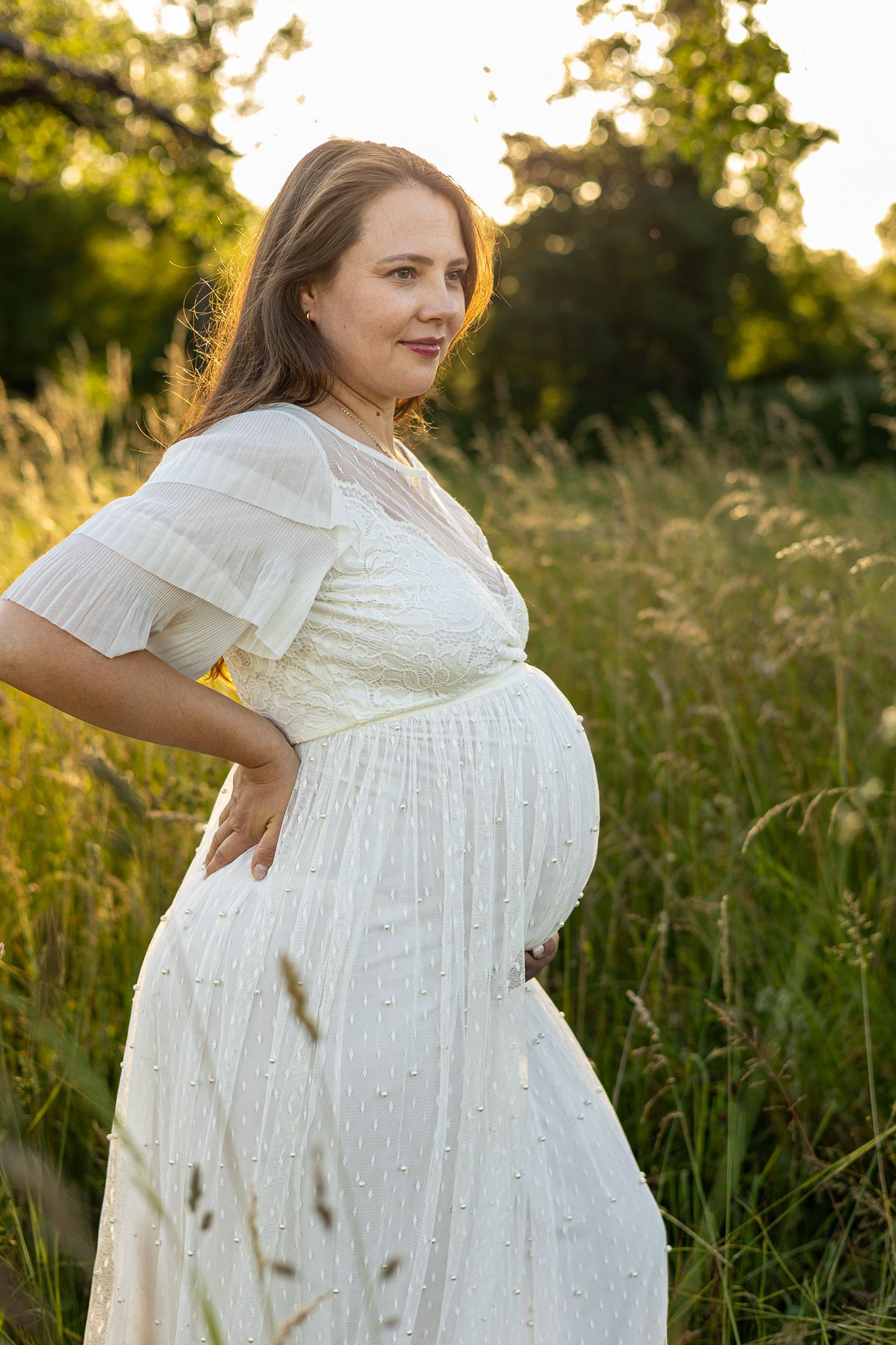 Pregnant woman in white lace dress standing in golden hour sunlight among tall green grass, cradling her baby bump.
