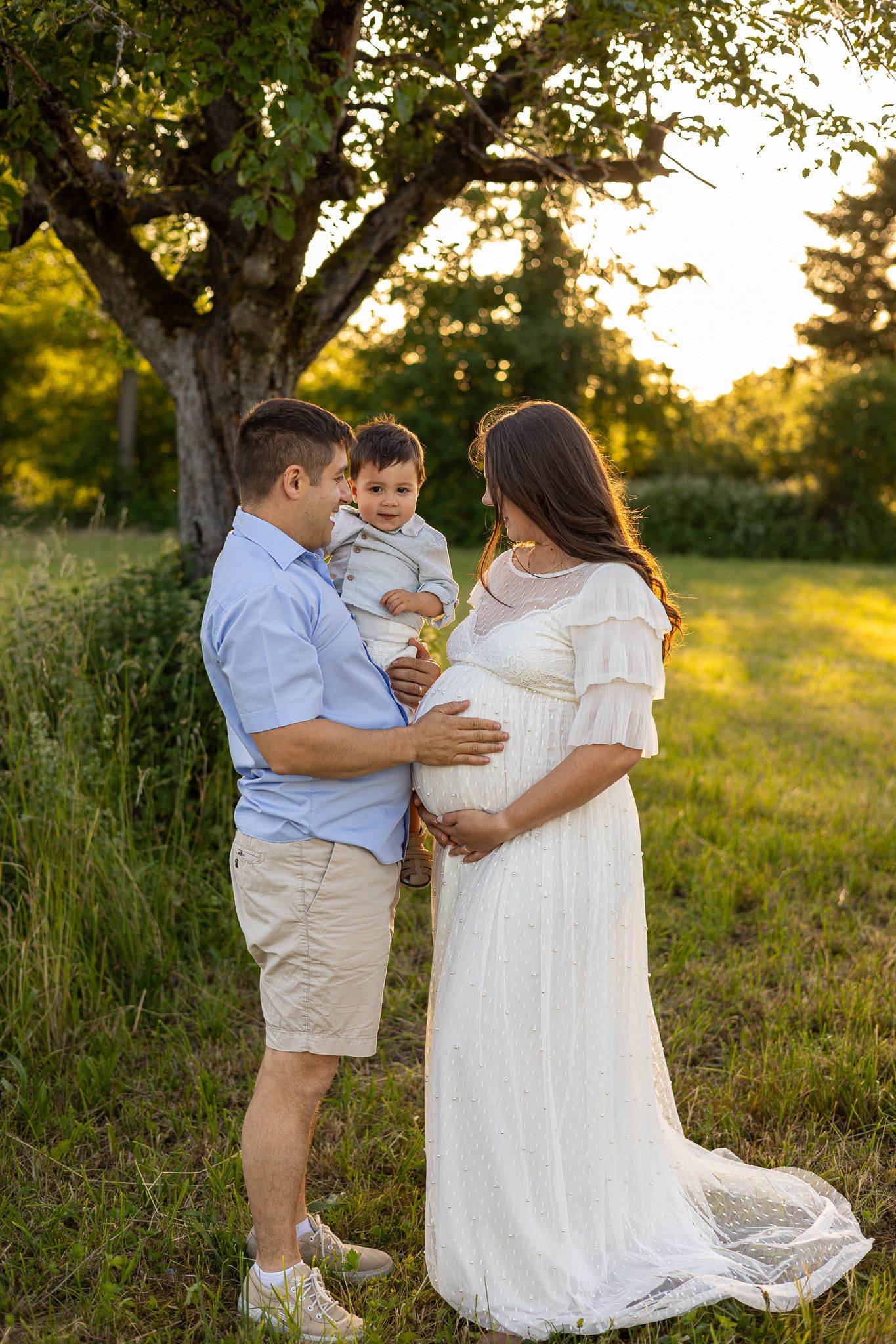 Pregnant woman in white lace gown with husband holding toddler son outdoors at golden hour sunset.