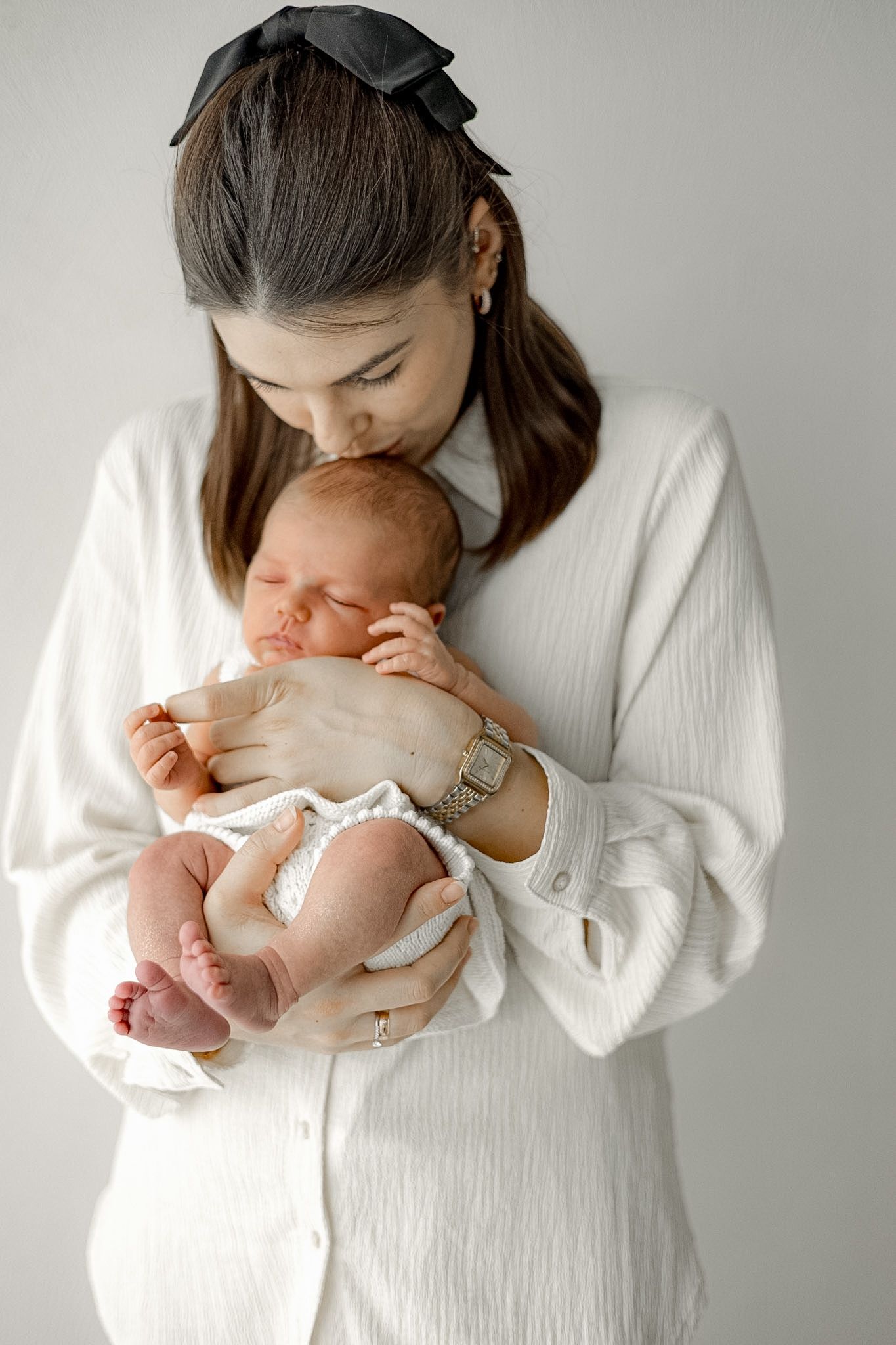 A mother tenderly kisses her sleeping newborn baby while cradling it in her arms, dressed in white.