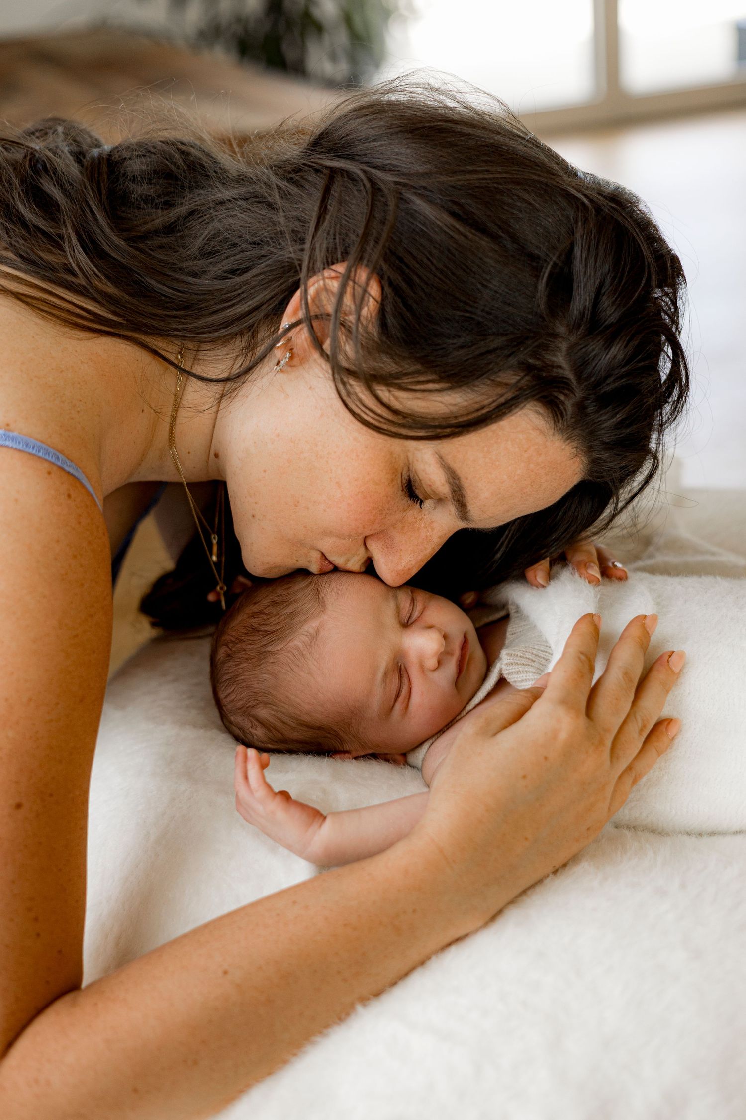 A loving mother gently kisses her sleeping newborn baby on a soft white blanket.