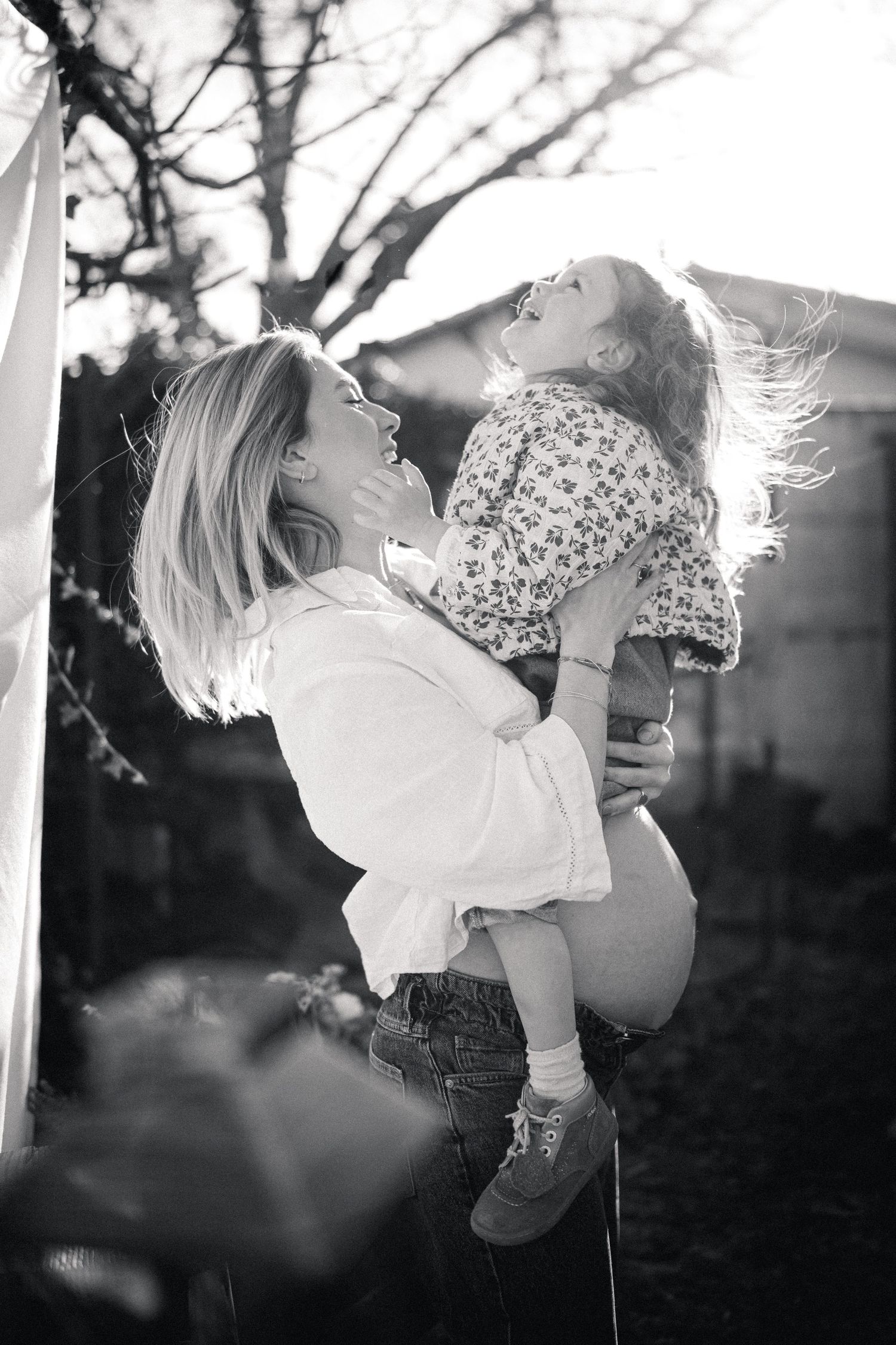 Black and white photo of a mother lifting her laughing toddler daughter in a sunny backyard.