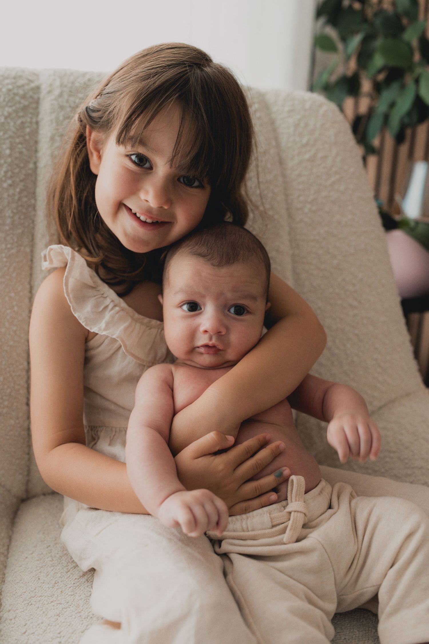 A smiling young girl proudly holds her newborn sibling in a cozy white armchair at home.