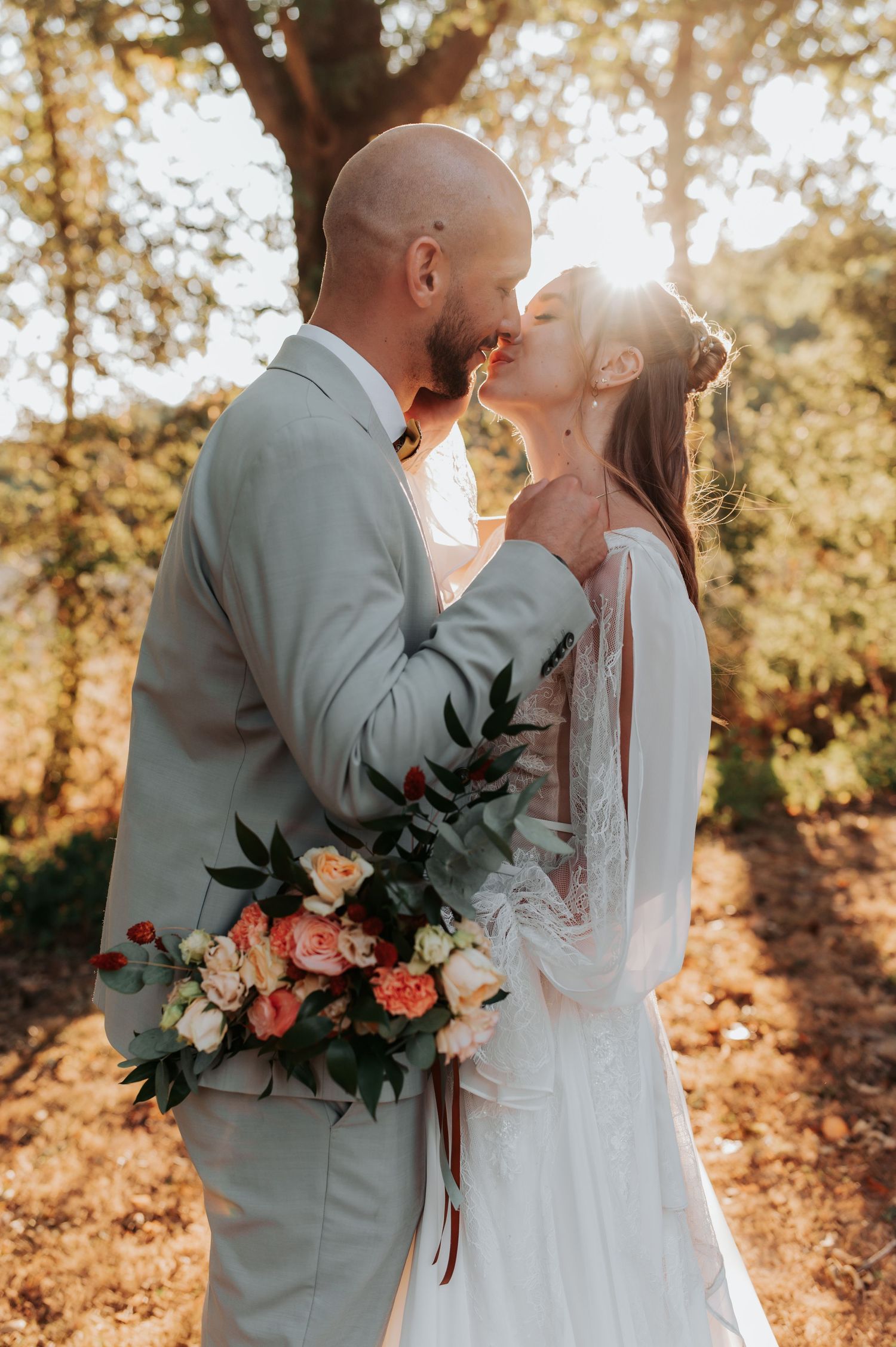 A bride and groom share a romantic kiss outdoors, with golden sunlight and a colorful bridal bouquet.