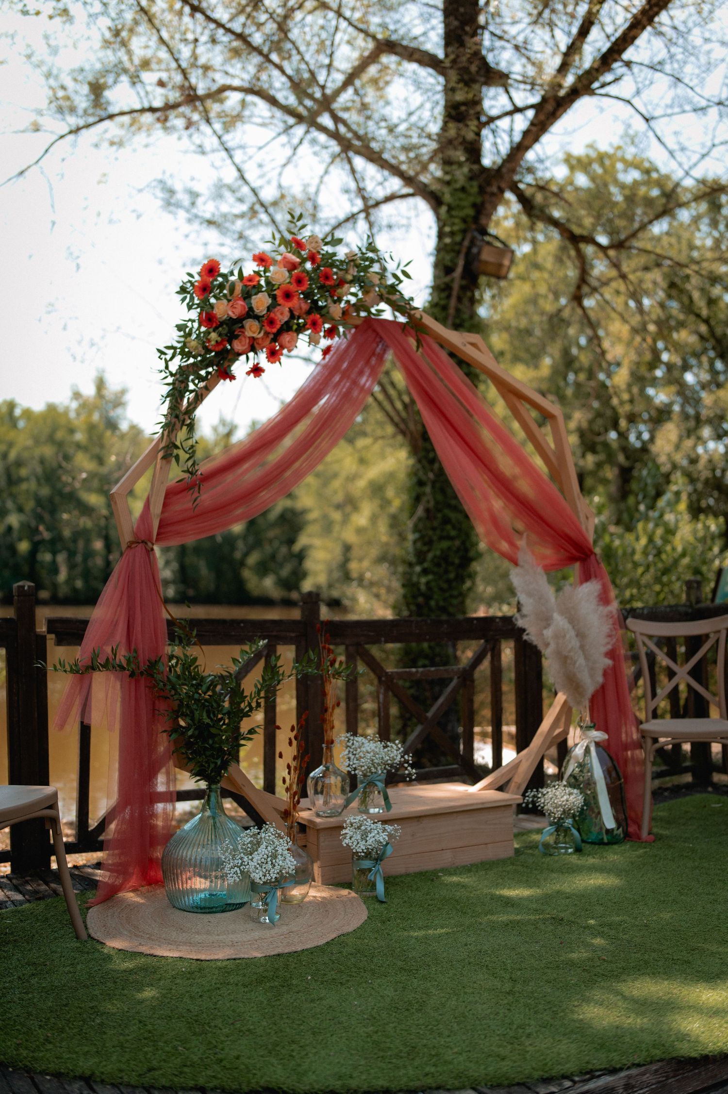 Outdoor wedding arch draped in coral tulle with floral arrangements, set on green grass with decorative vases nearby.