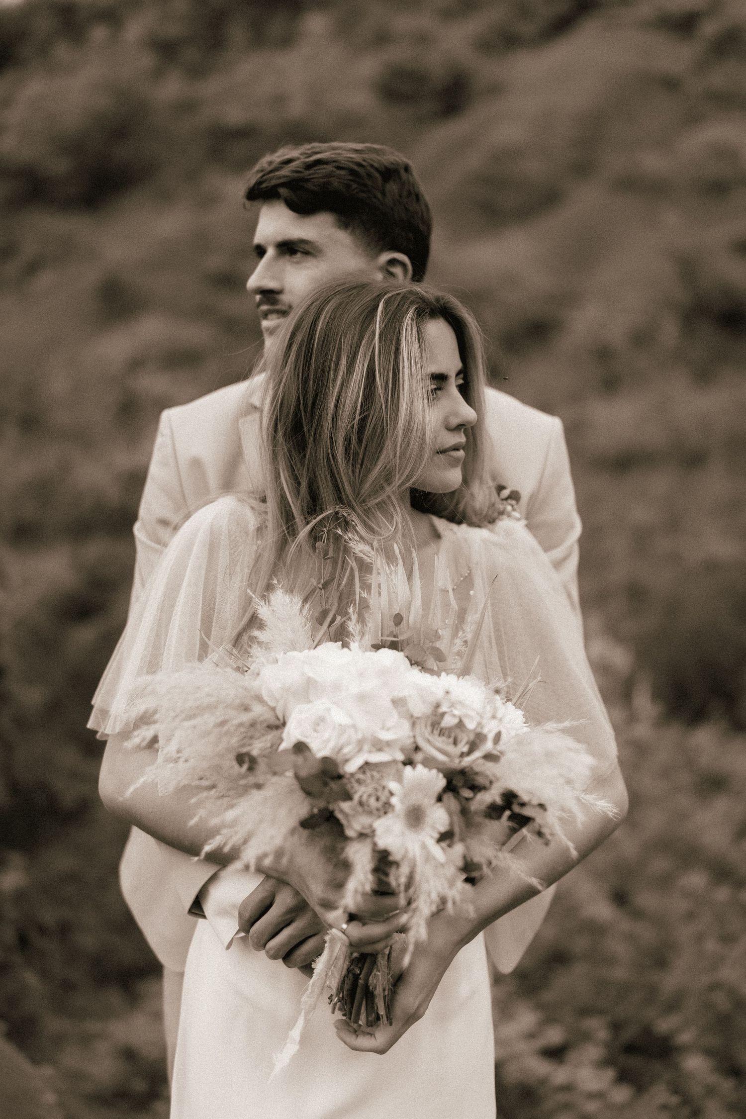 Bride and groom embrace in a romantic sepia-toned outdoor wedding portrait with a floral bouquet.