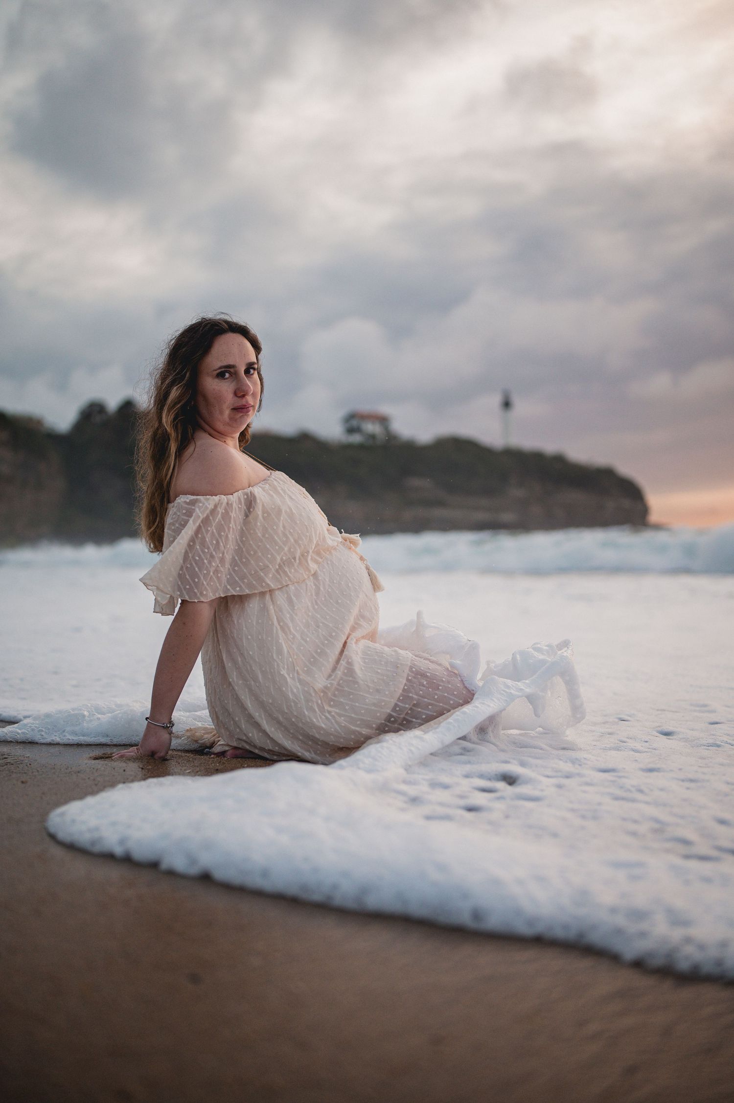 Pregnant woman in white lace dress sitting on beach shore with waves and lighthouse at sunset.