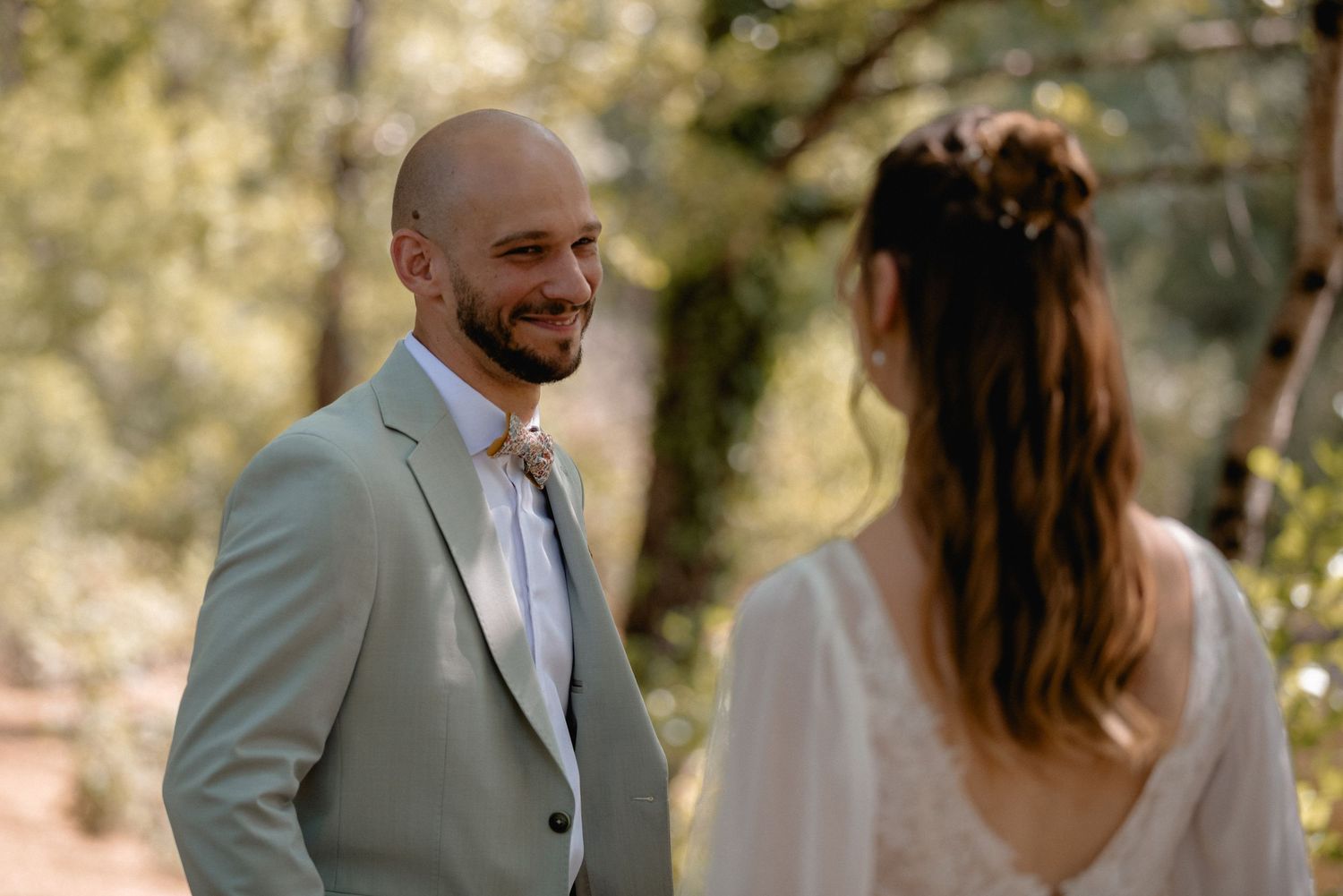 Groom in sage green suit smiling at bride during outdoor wedding ceremony among trees.