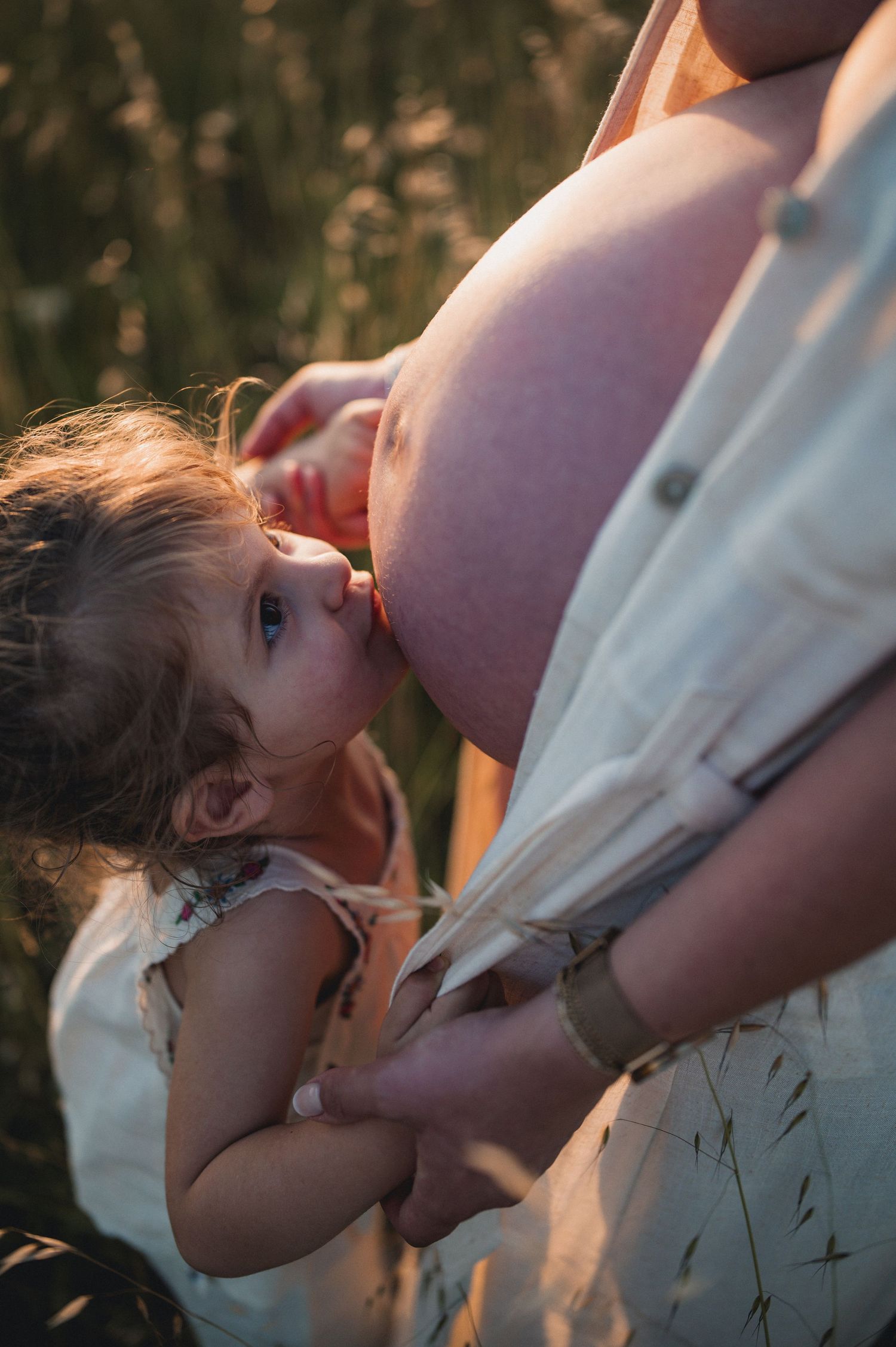 Young girl resting head against pregnant mothers bare belly in golden sunlit field.