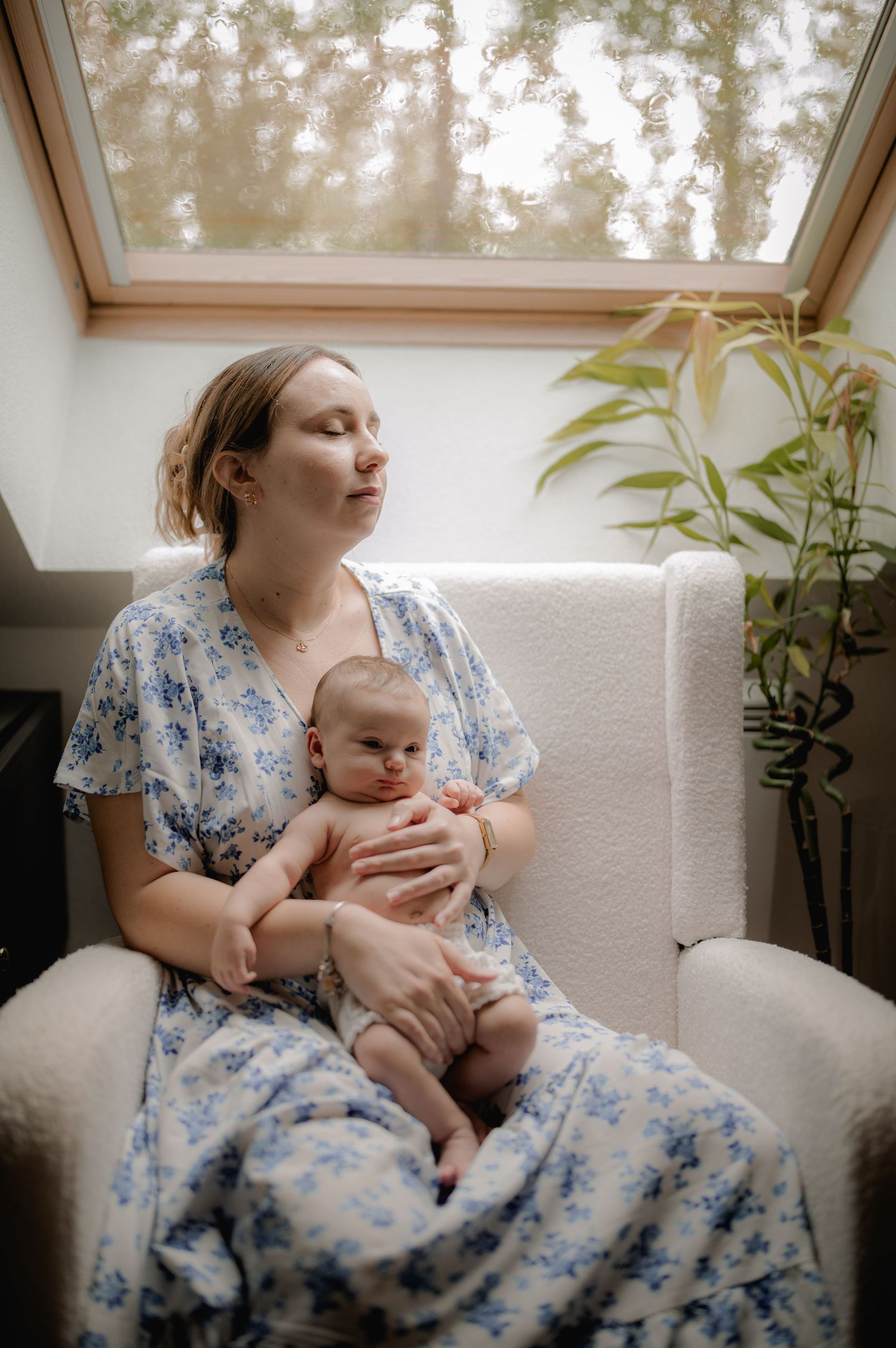 New mother in floral blue dress sitting in white armchair holding newborn baby by window.