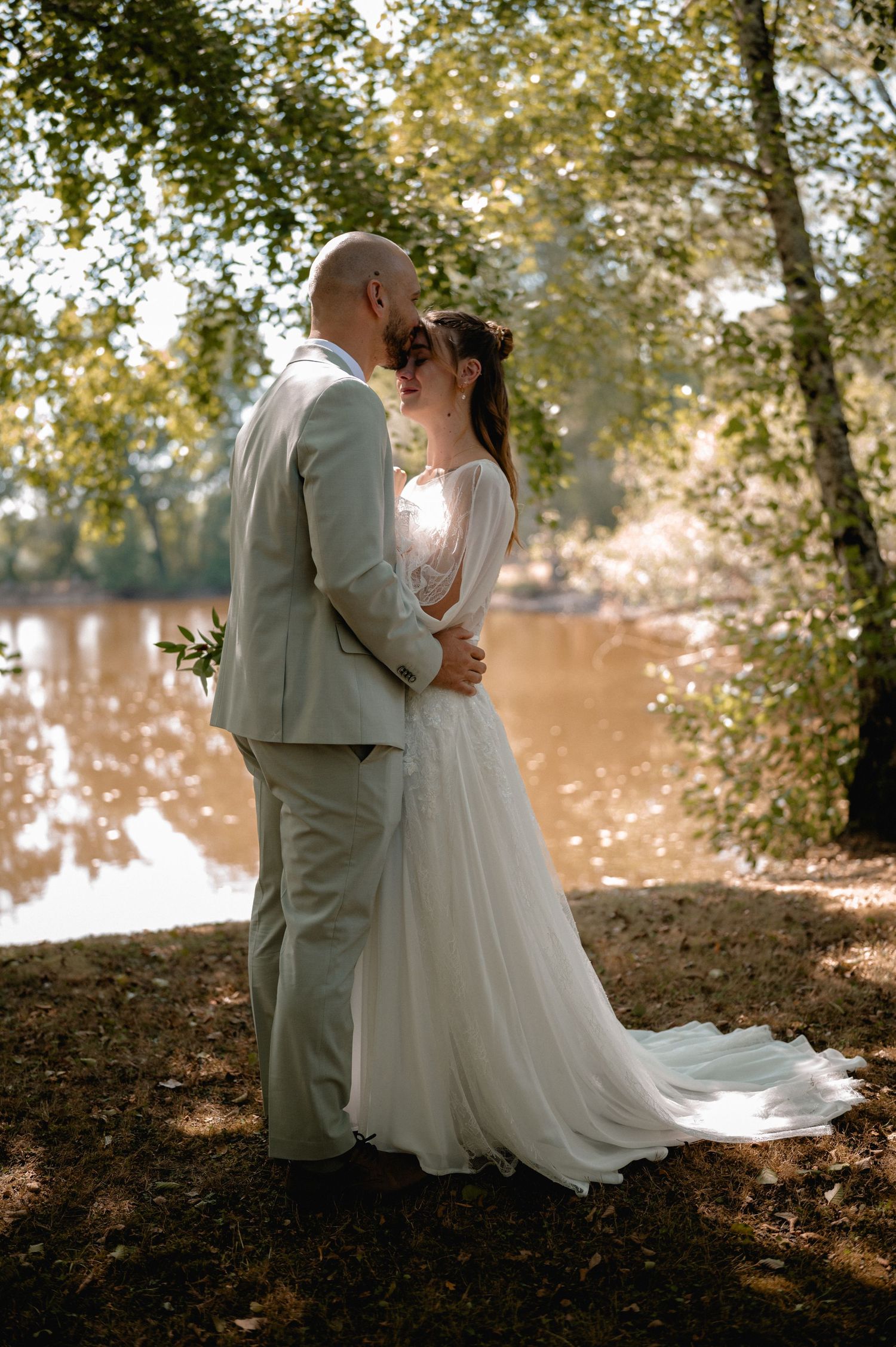 Bride and groom share a romantic moment by a sunlit lake, surrounded by trees, as she wears a flowing white gown.