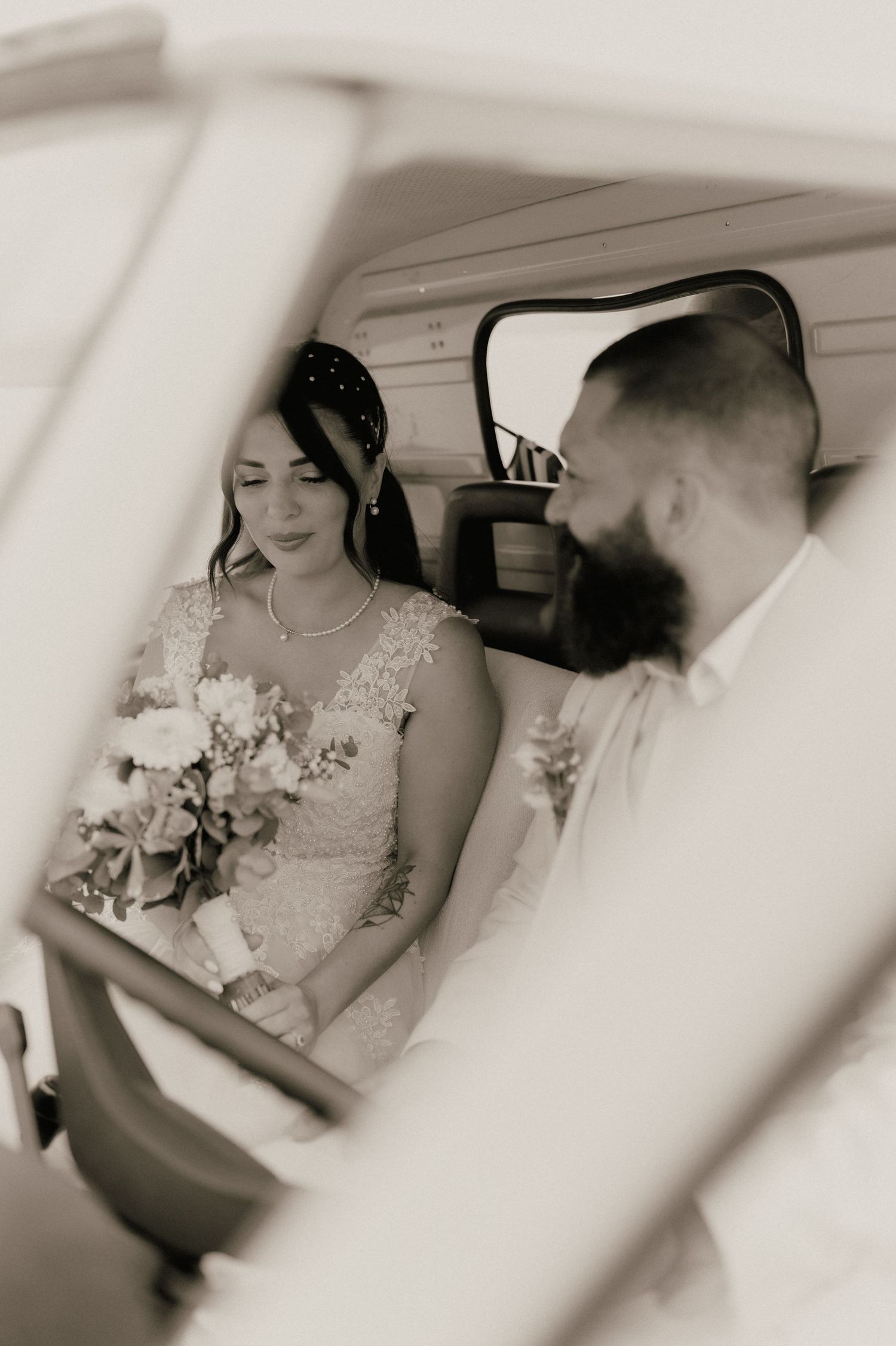 Black and white photo of a bride holding a bouquet and groom with beard sitting together inside a vintage vehicle on wedding day.