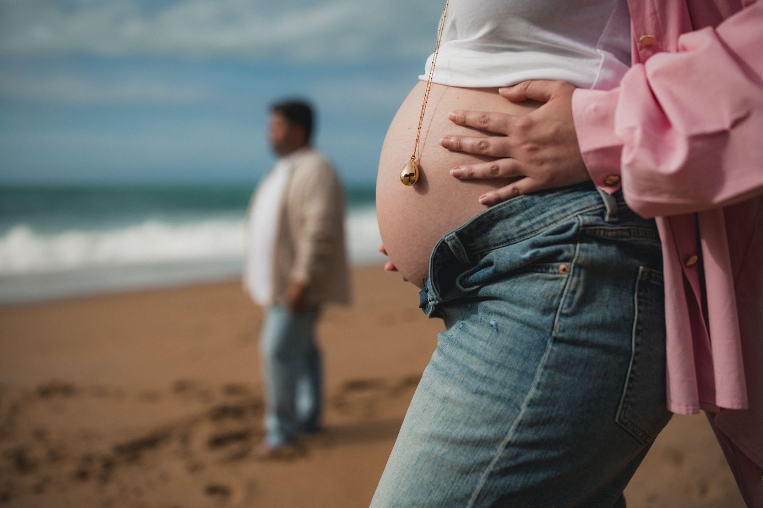 Close-up of pregnant belly in jeans with pink shirt at beach, man standing in background by ocean.