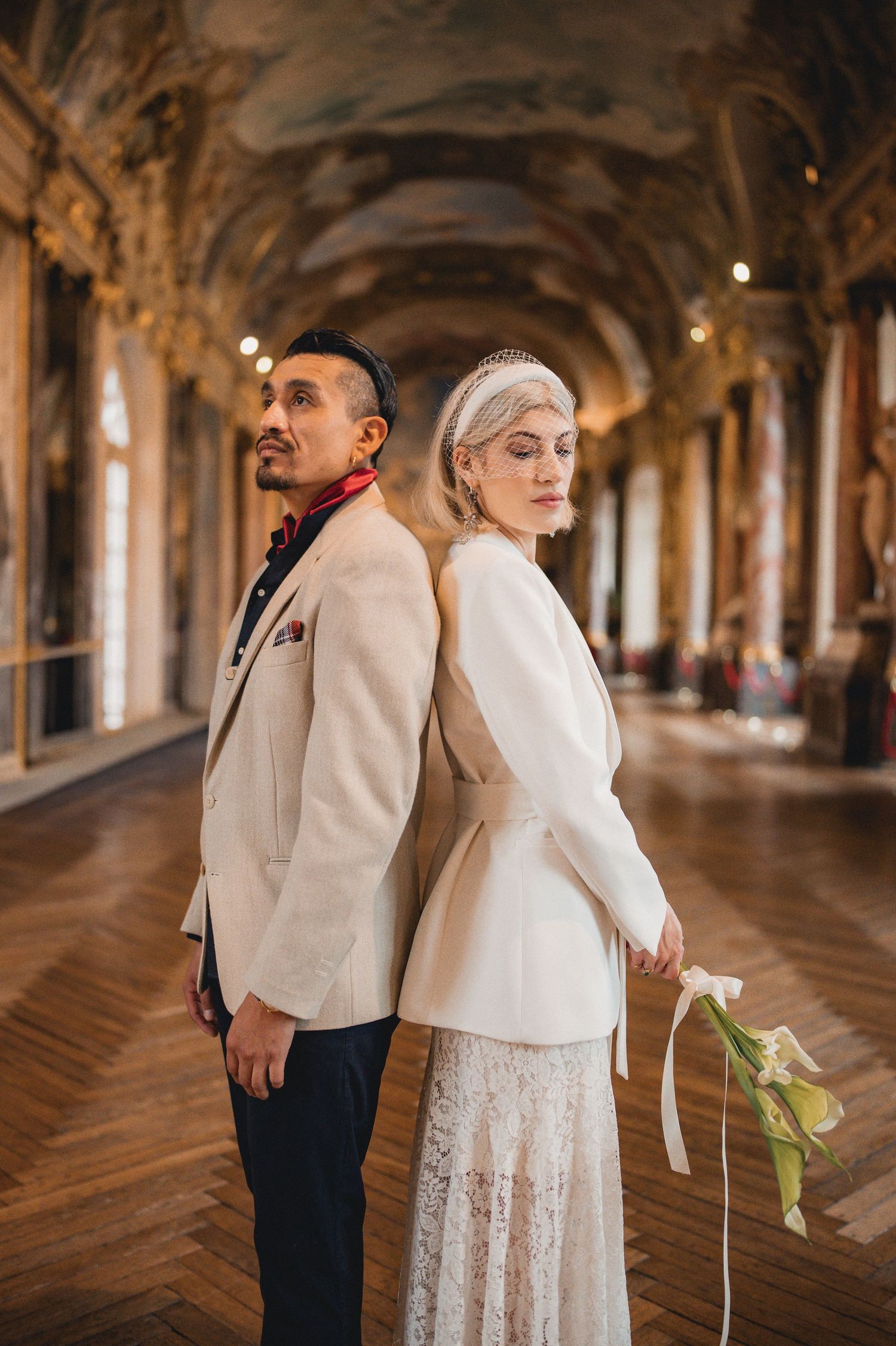 Elegant couple posing back-to-back in ornate Versailles hall, bride in white lace dress holding flowers, groom in cream blazer.