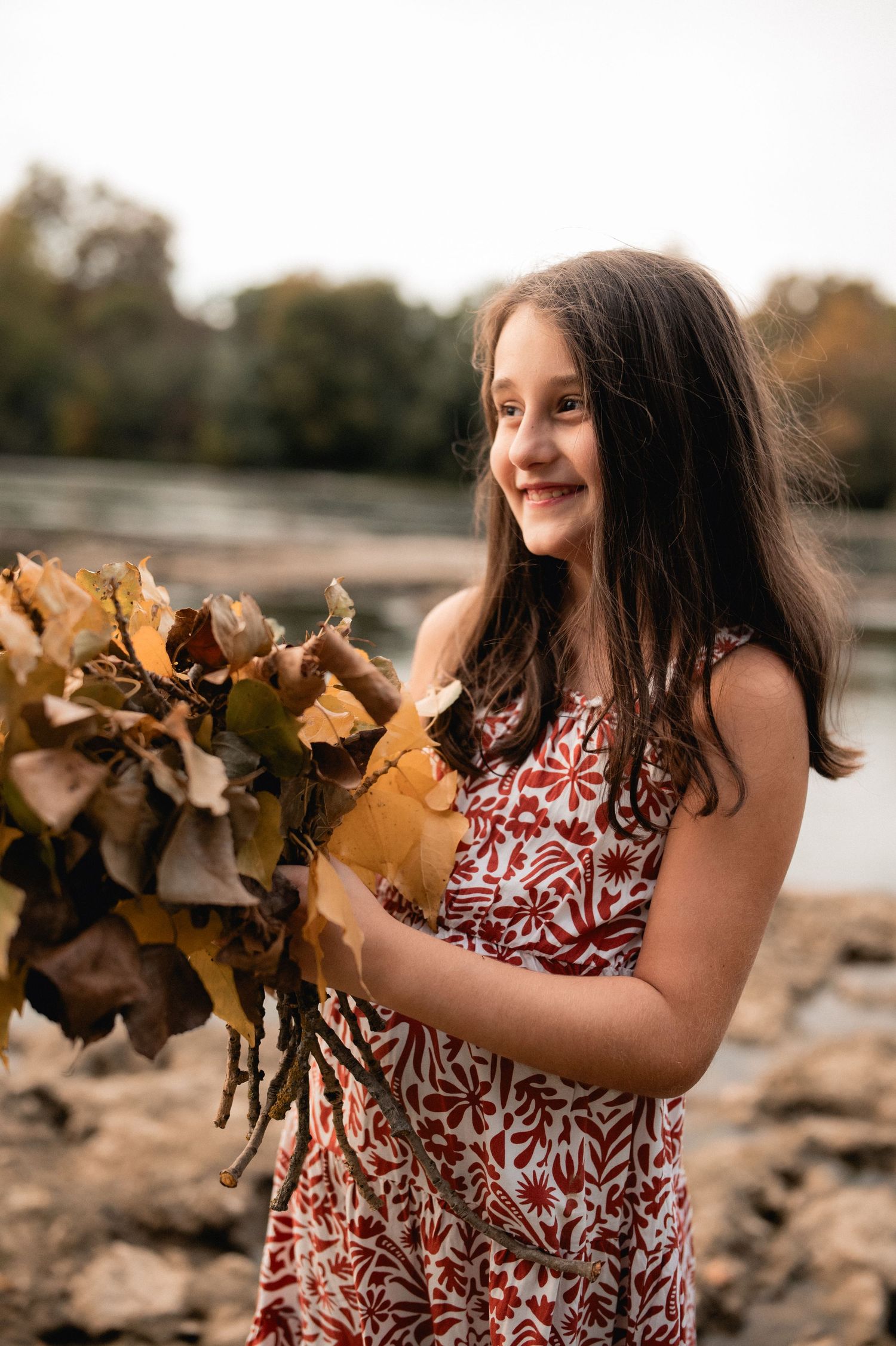 Smiling young girl holding a bouquet of autumn leaves by a river in a floral red dress.