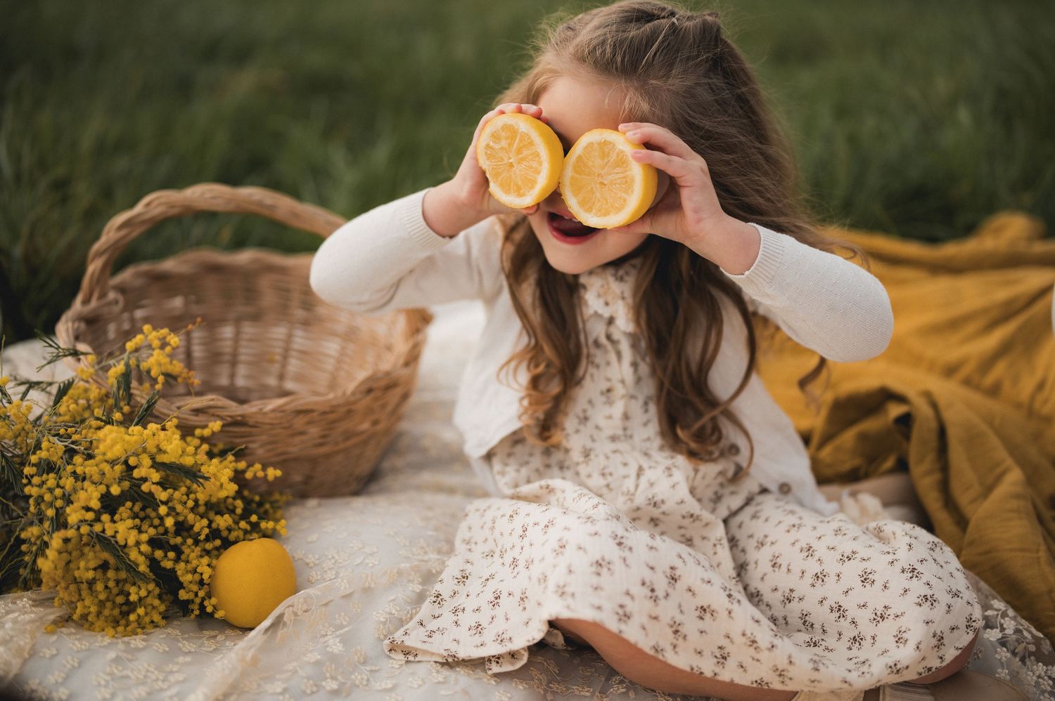 Young girl sitting outdoors holding lemon halves over eyes near wicker basket with flowers.