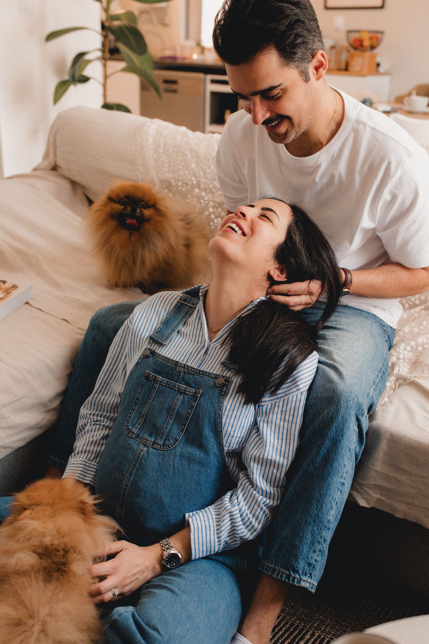 Smiling pregnant woman in denim overalls leaning on partner with fluffy Pomeranian dog indoors.