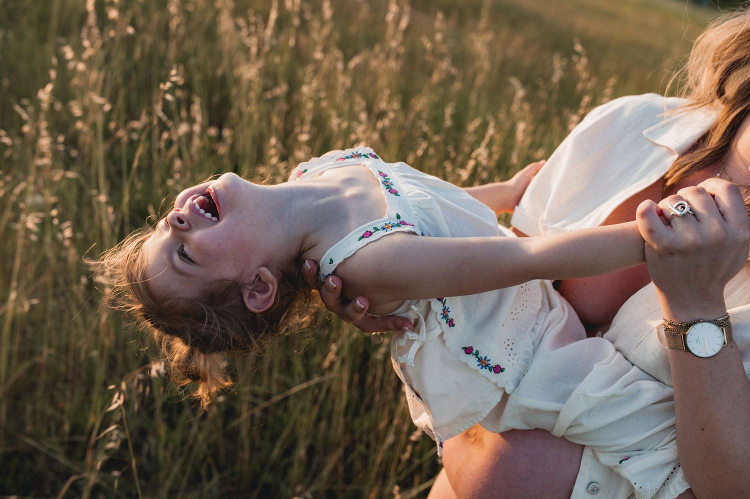 A laughing child is held and spun by an adult in a sunlit golden grass field.