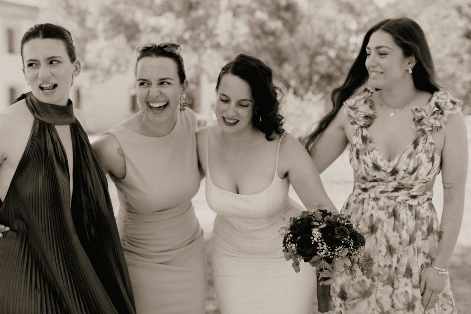 Black and white photo of a bride in white dress laughing with three bridesmaids outdoors at a wedding celebration.