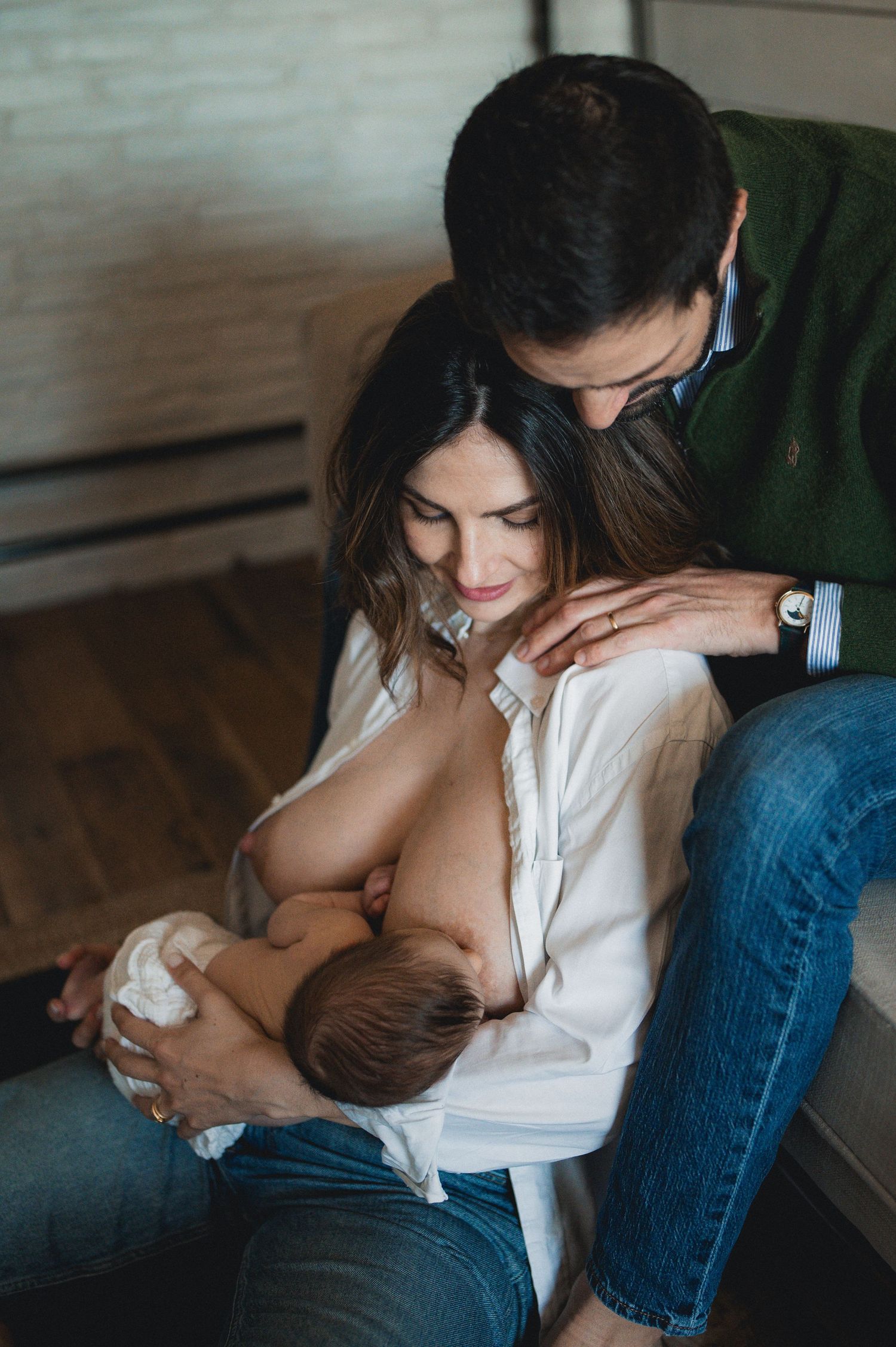 Mother breastfeeding newborn baby while father lovingly embraces her during an indoor lifestyle newborn session.