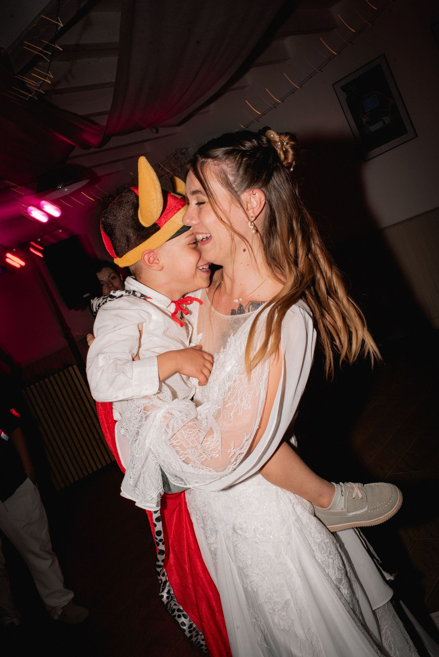 A bride in a white lace dress holds a child in a devil costume, both laughing joyfully on a wedding dance floor.