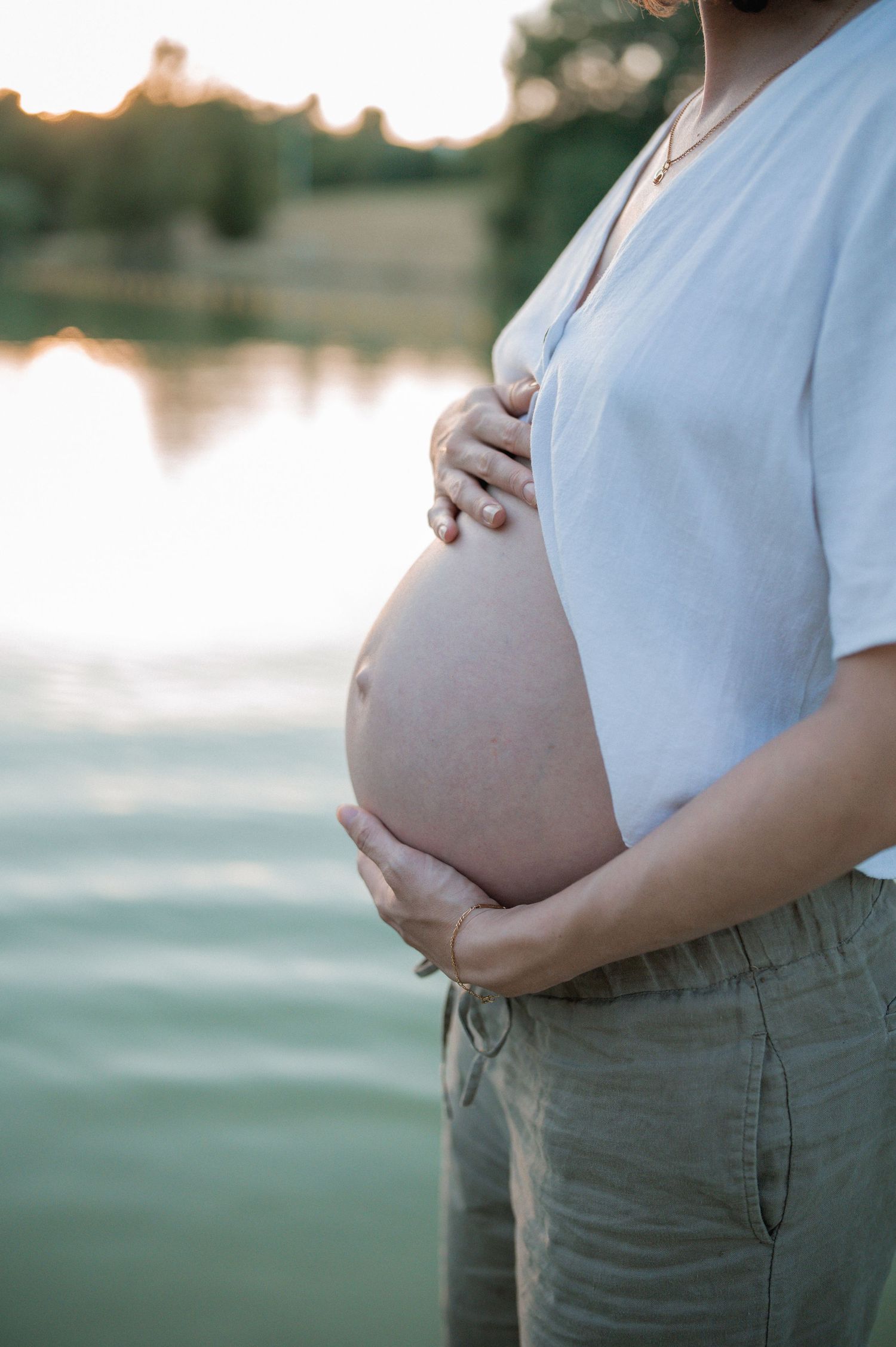 Pregnant woman in white shirt cradling bare baby bump with lake and trees in soft sunset background.