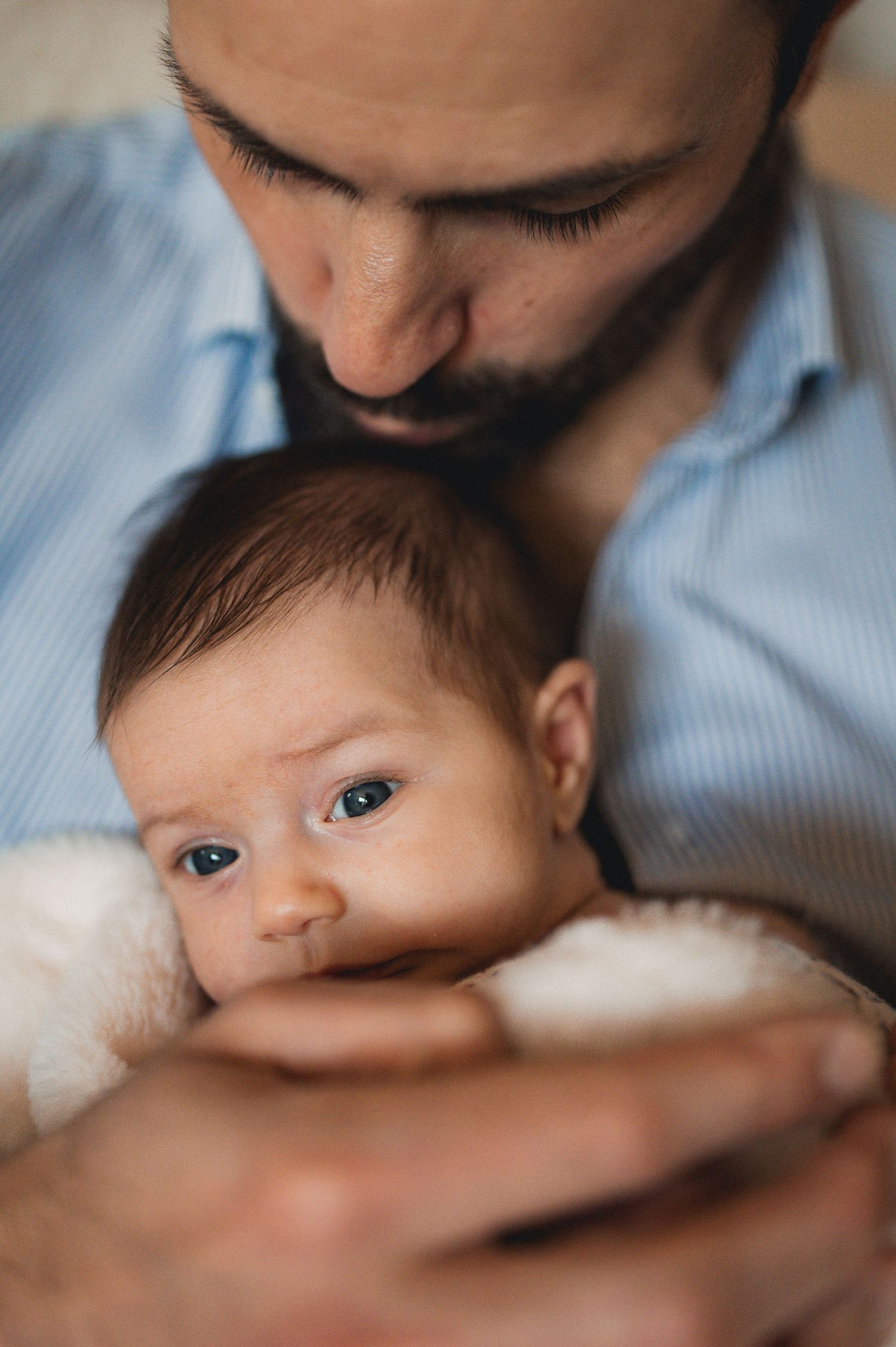 Bearded father in blue shirt cradling alert newborn baby wrapped in white blanket.