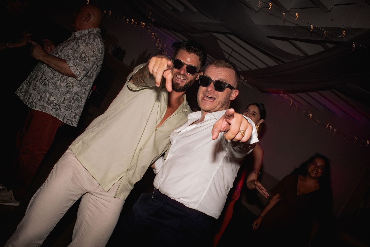 Two men in light-colored outfits pointing at camera while dancing at a party event.