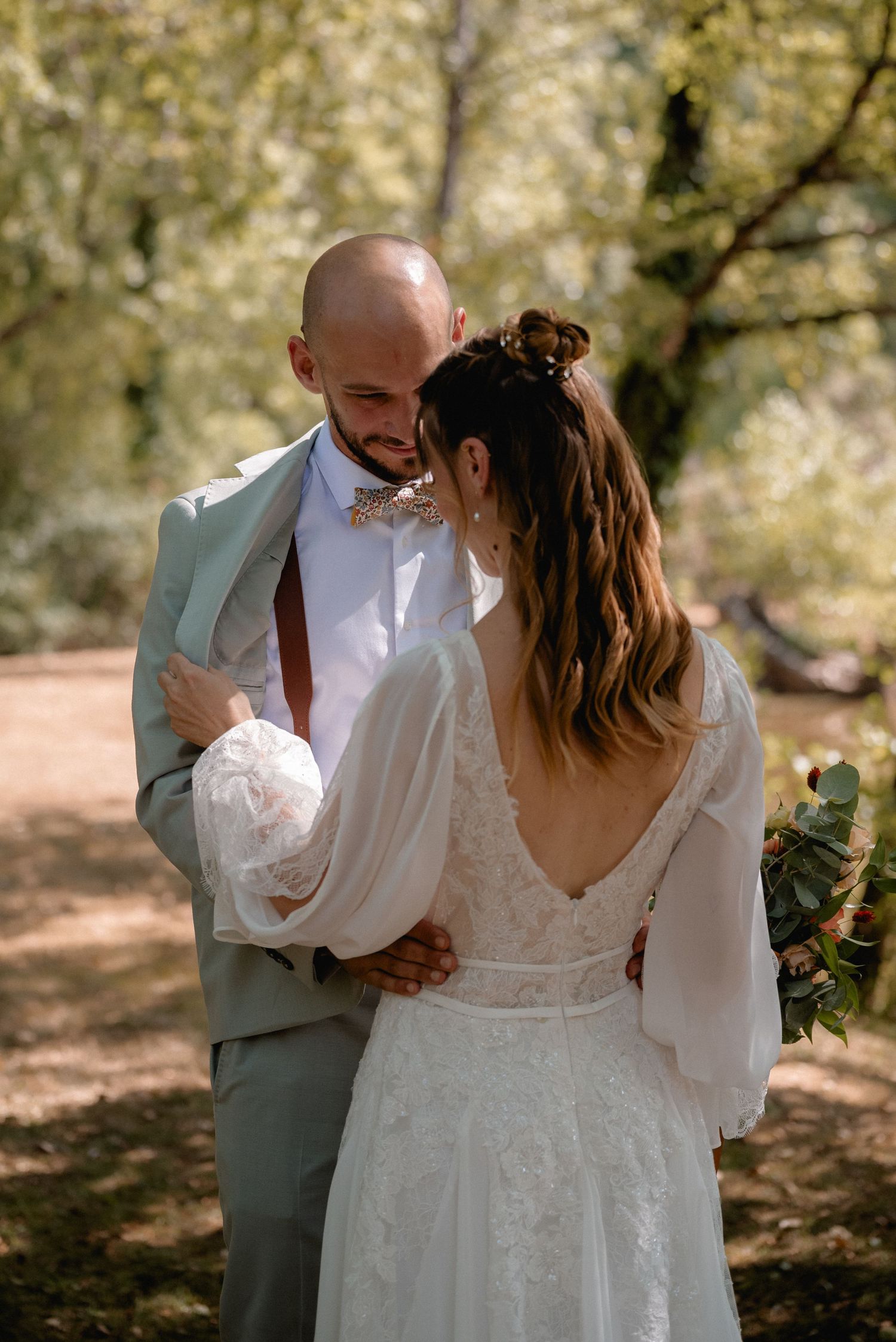 A bride and groom share a tender moment in a sunlit forest, she in lace gown, he in sage suit with bow tie.