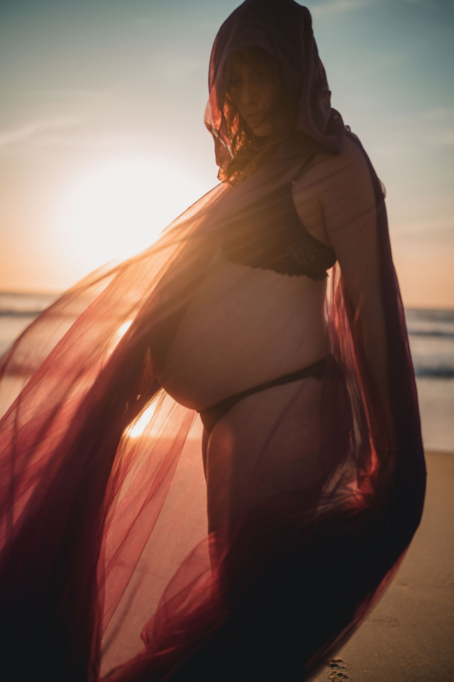 Pregnant woman in black bikini draped in flowing burgundy tulle fabric backlit by golden sunset on beach.
